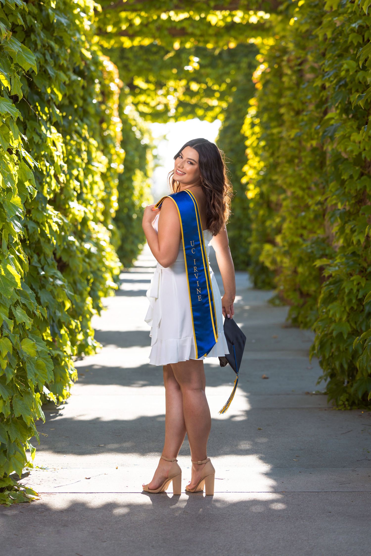 Graduate wearing white dress and blue stole poses on pathway surrounded by lush green foliage.