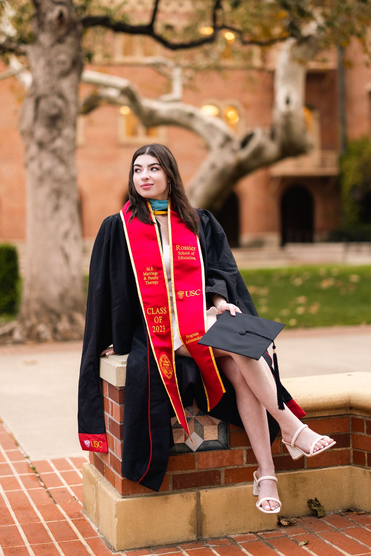 Graduate in cap and gown sitting on brick wall with red graduation sash.