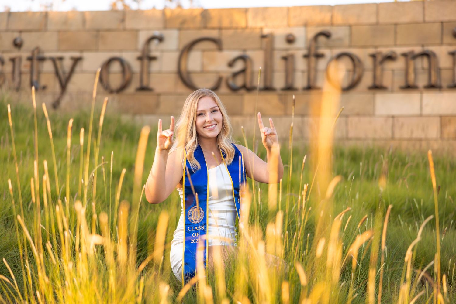 Graduate celebrates by posing in front of University of California sign surrounded by golden grass.
