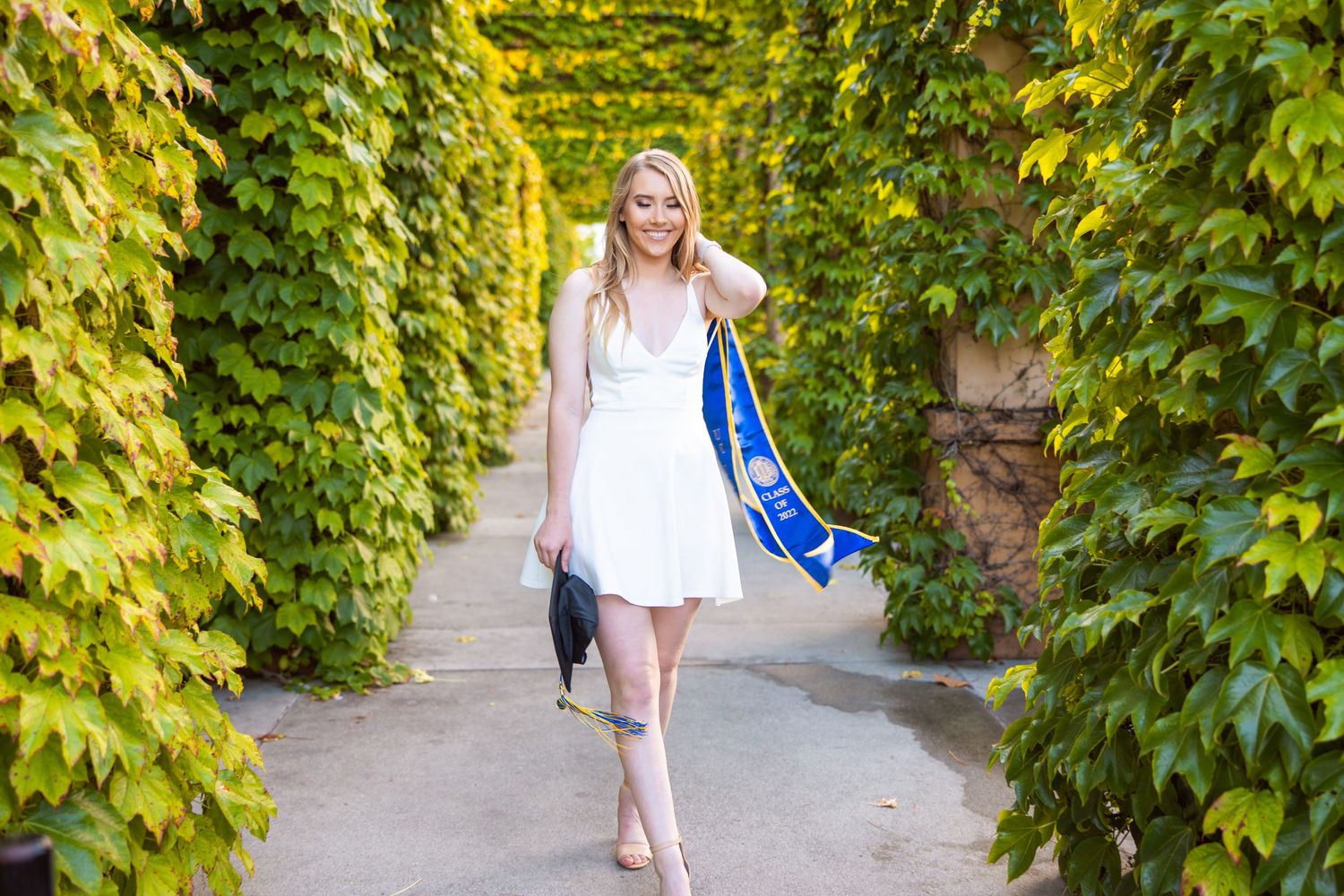 Graduate wearing white dress and blue graduation stole walking through ivy covered pathway at UC Irvine.