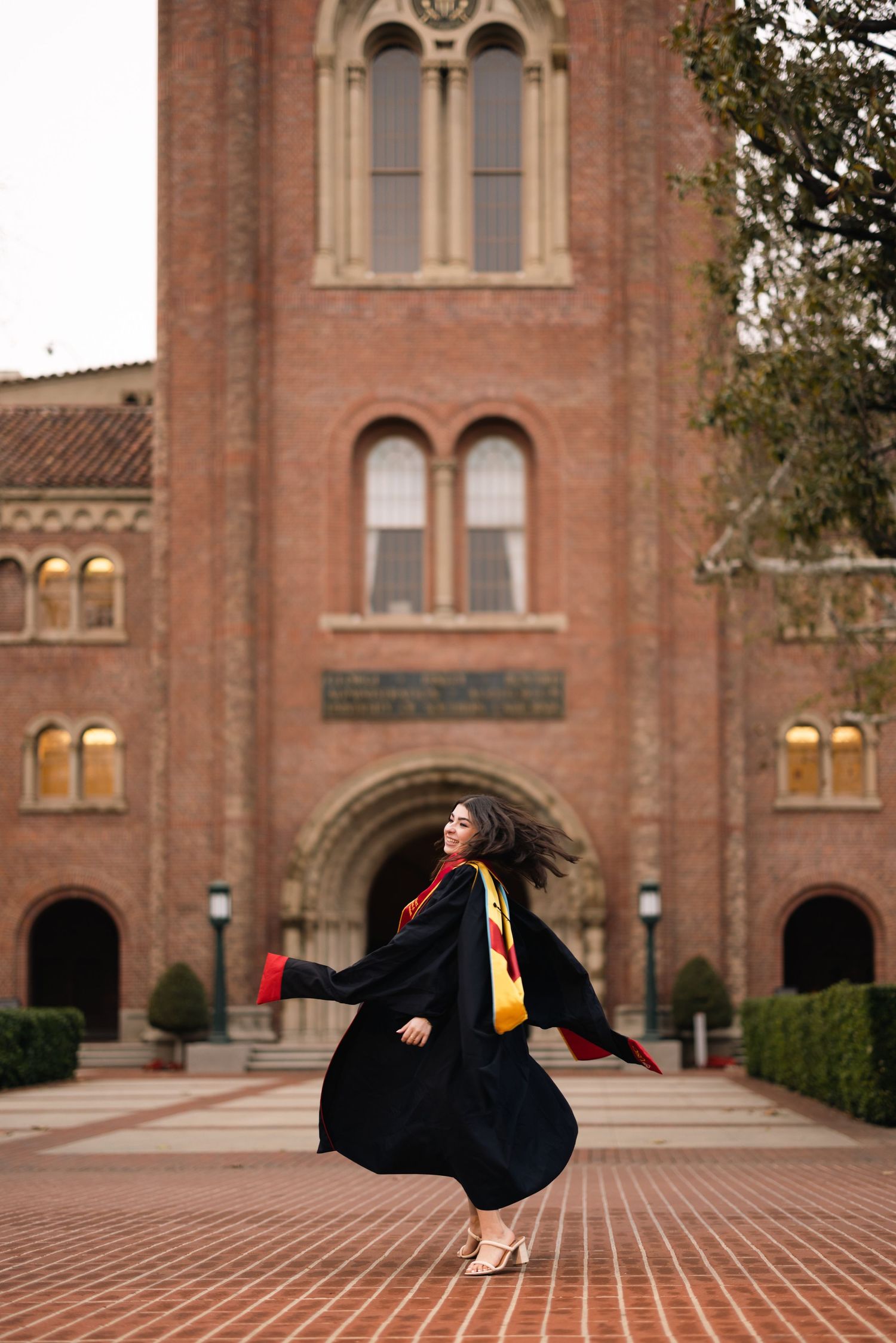 Graduate twirling in black graduation gown in front of historic university building.
