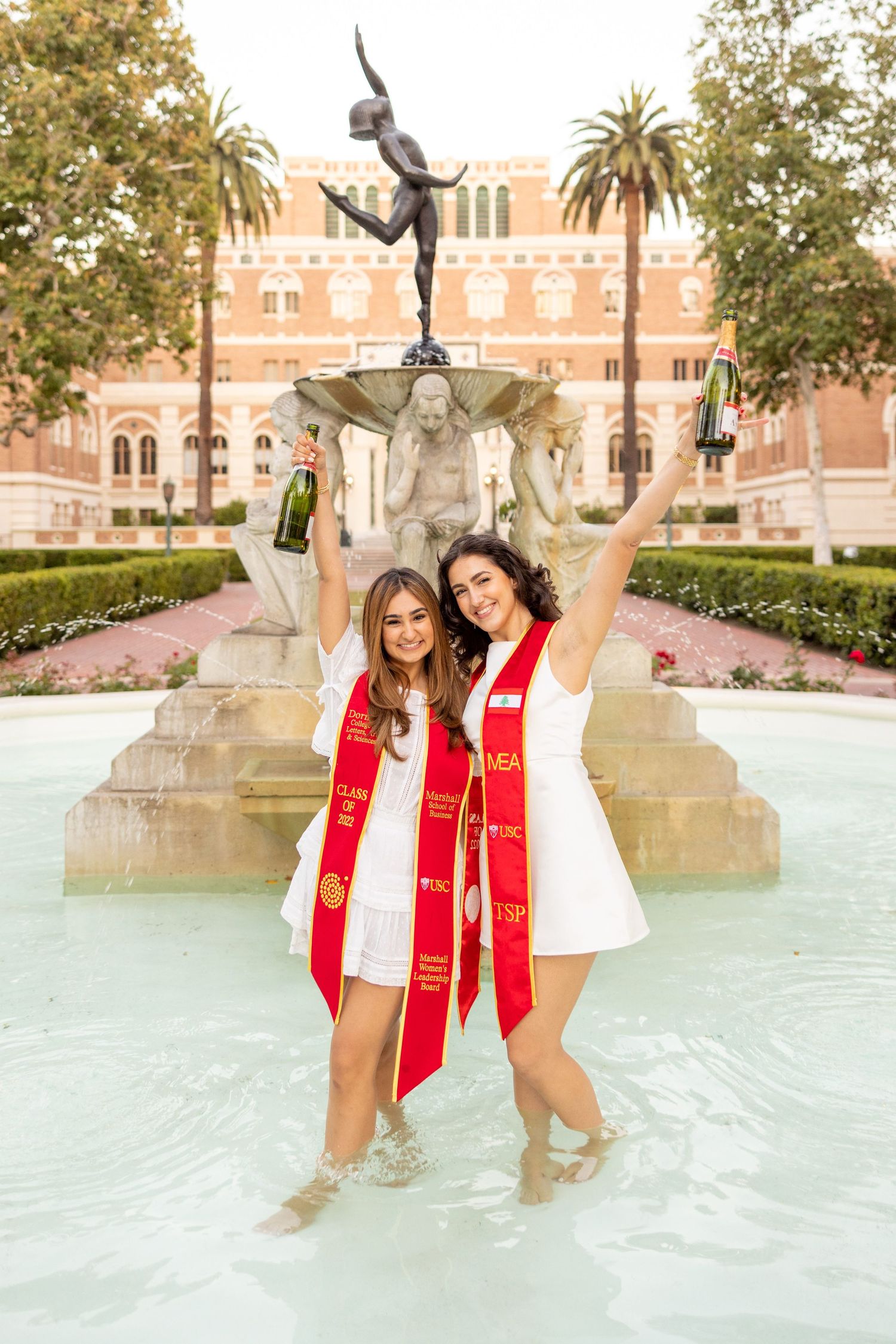 Two graduates celebrate in fountain water wearing red USC stoles.