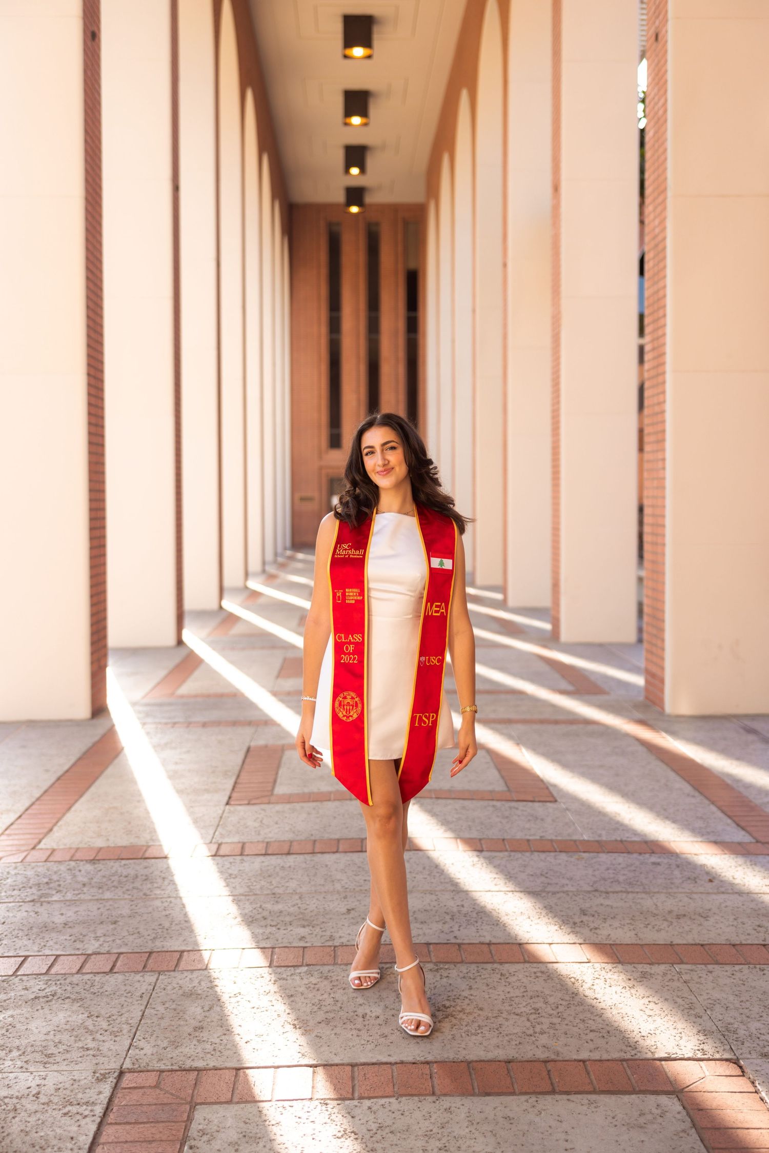Graduate in red stole poses in sunlit campus corridor with architectural columns.
