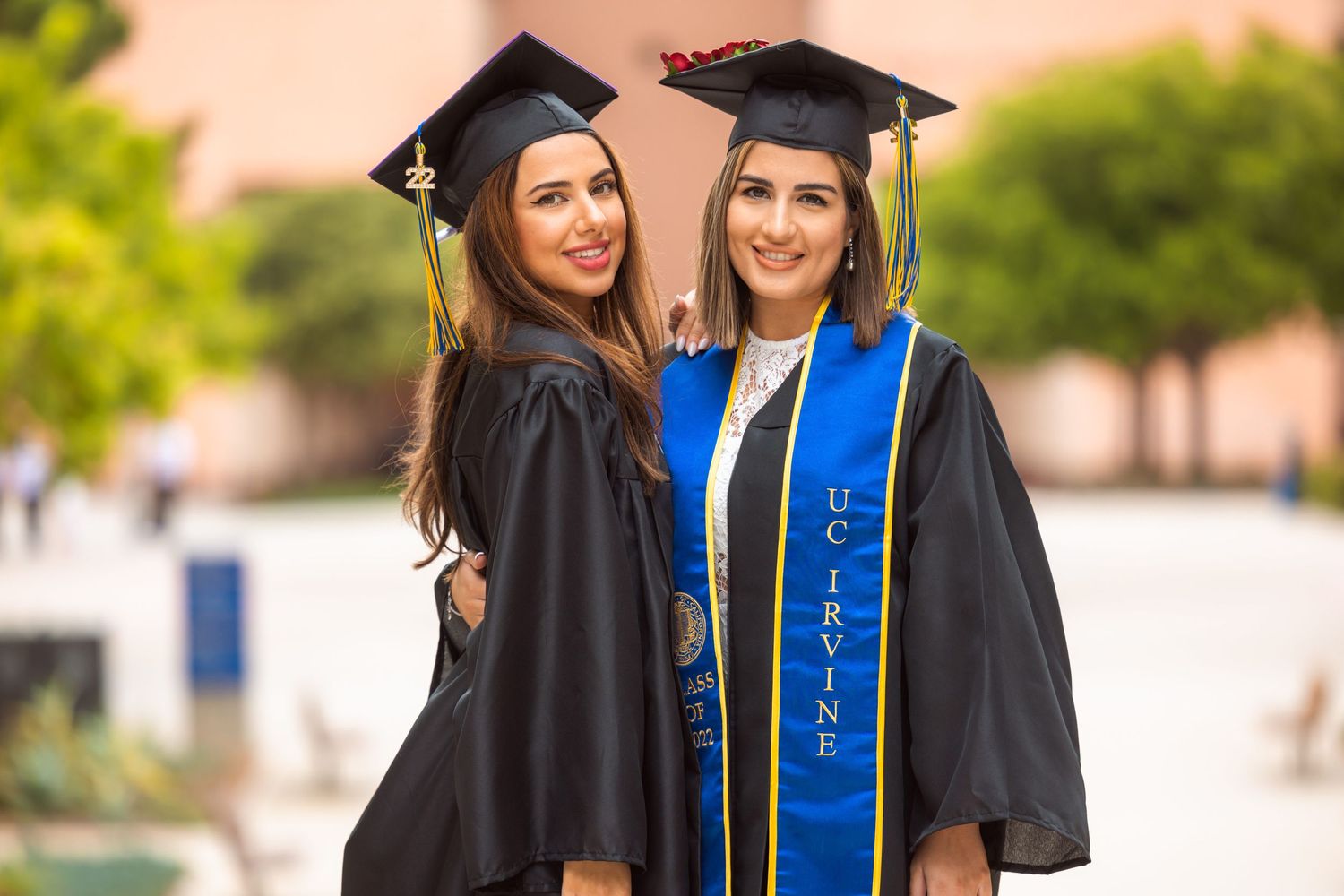 Two graduates in caps and gowns celebrate on campus wearing blue and gold stoles.