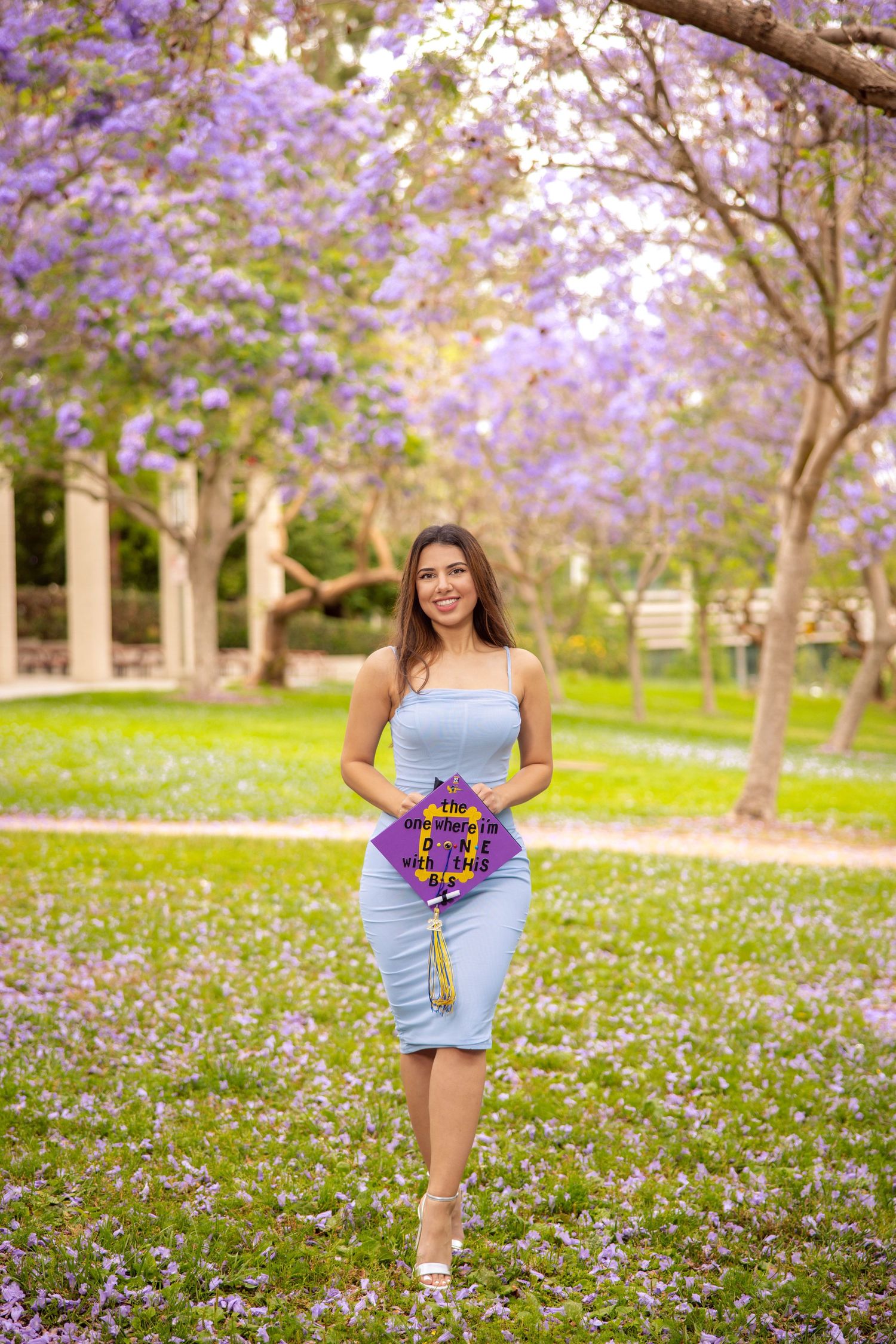 A graduate in a blue dress poses with flowers under blooming purple jacaranda trees.