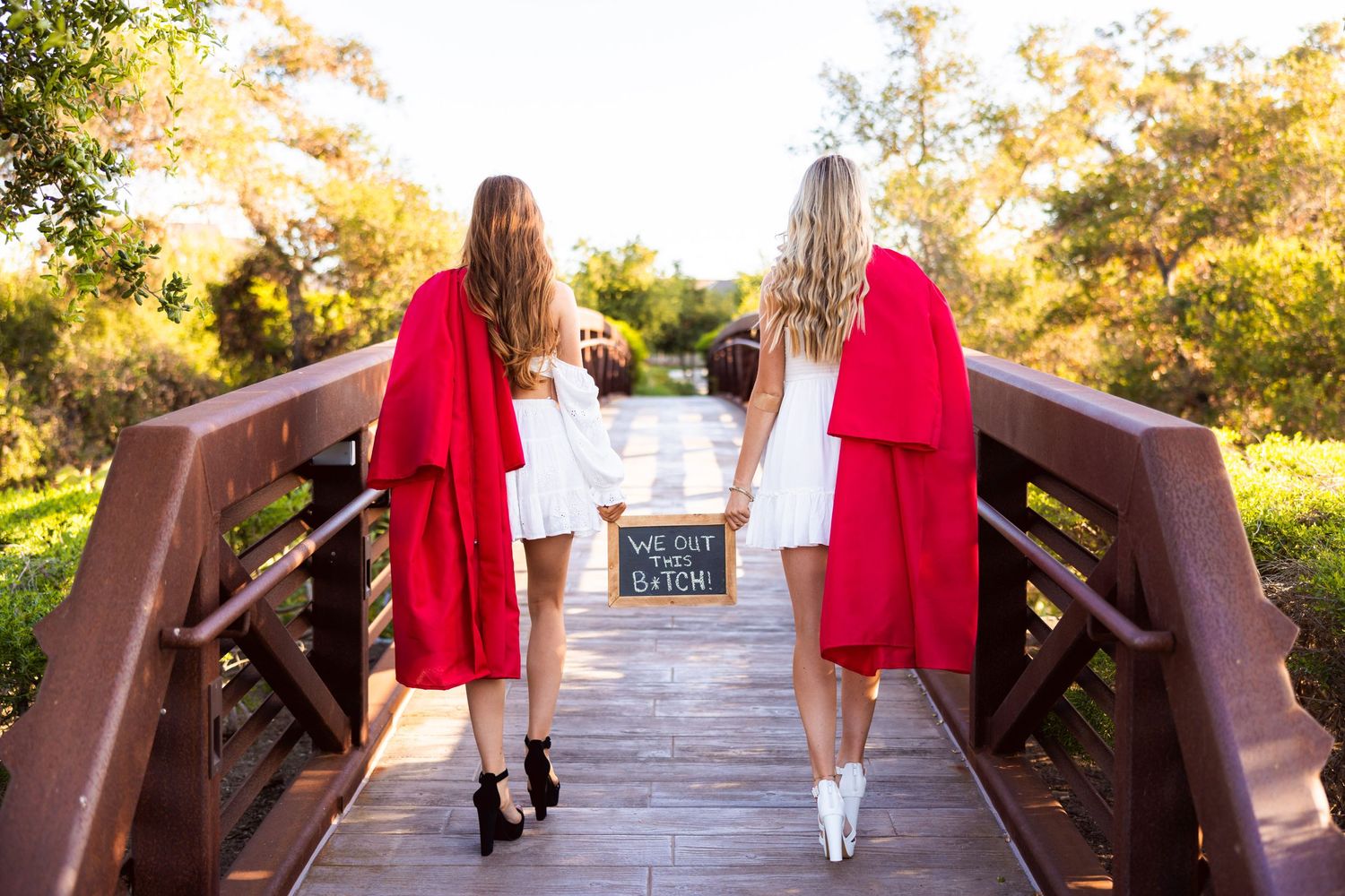 Two graduates in red gowns stand back-to-back on a wooden bridge during golden hour sunset.