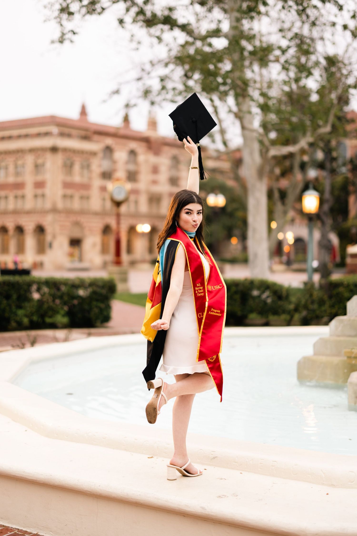 Graduate tossing graduation cap in celebratory pose by campus fountain at sunset.