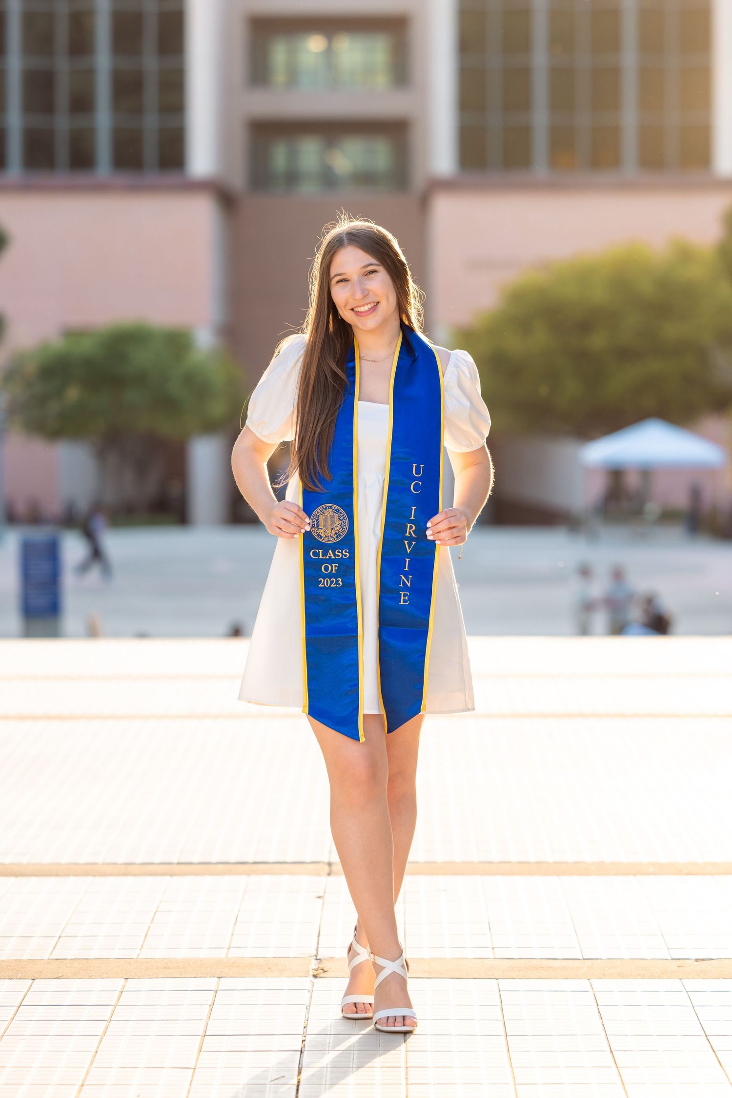 Graduate in white dress and blue graduation stole posing outdoors on campus.