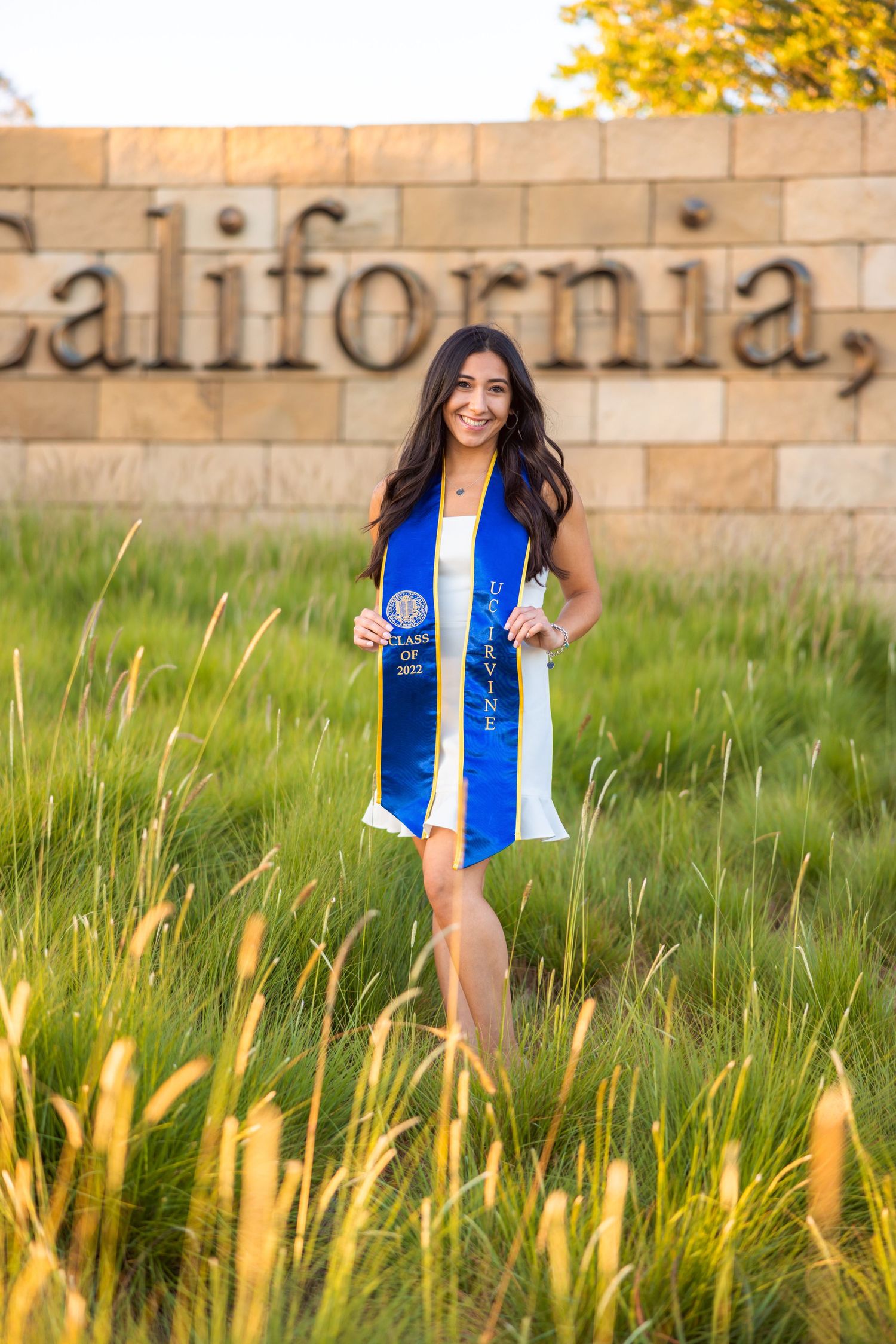 Graduate posing in white dress and blue stole by University of California sign with grass in foreground.
