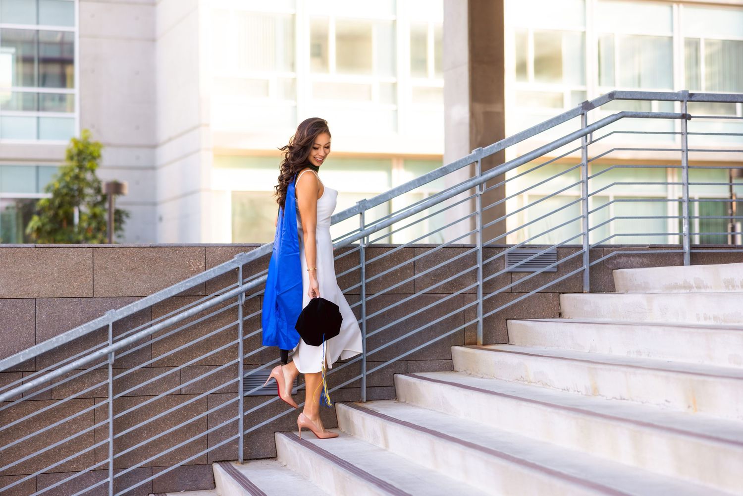 Graduate in blue dress descends modern concrete stairs while holding graduation cap.