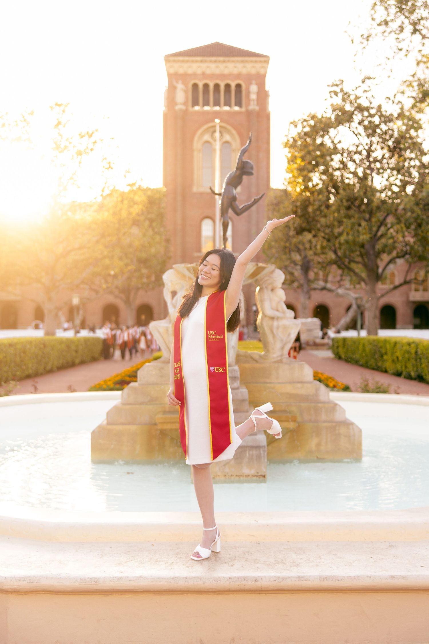 Graduate in red stole celebrates with arms raised in front of campus tower at sunset.