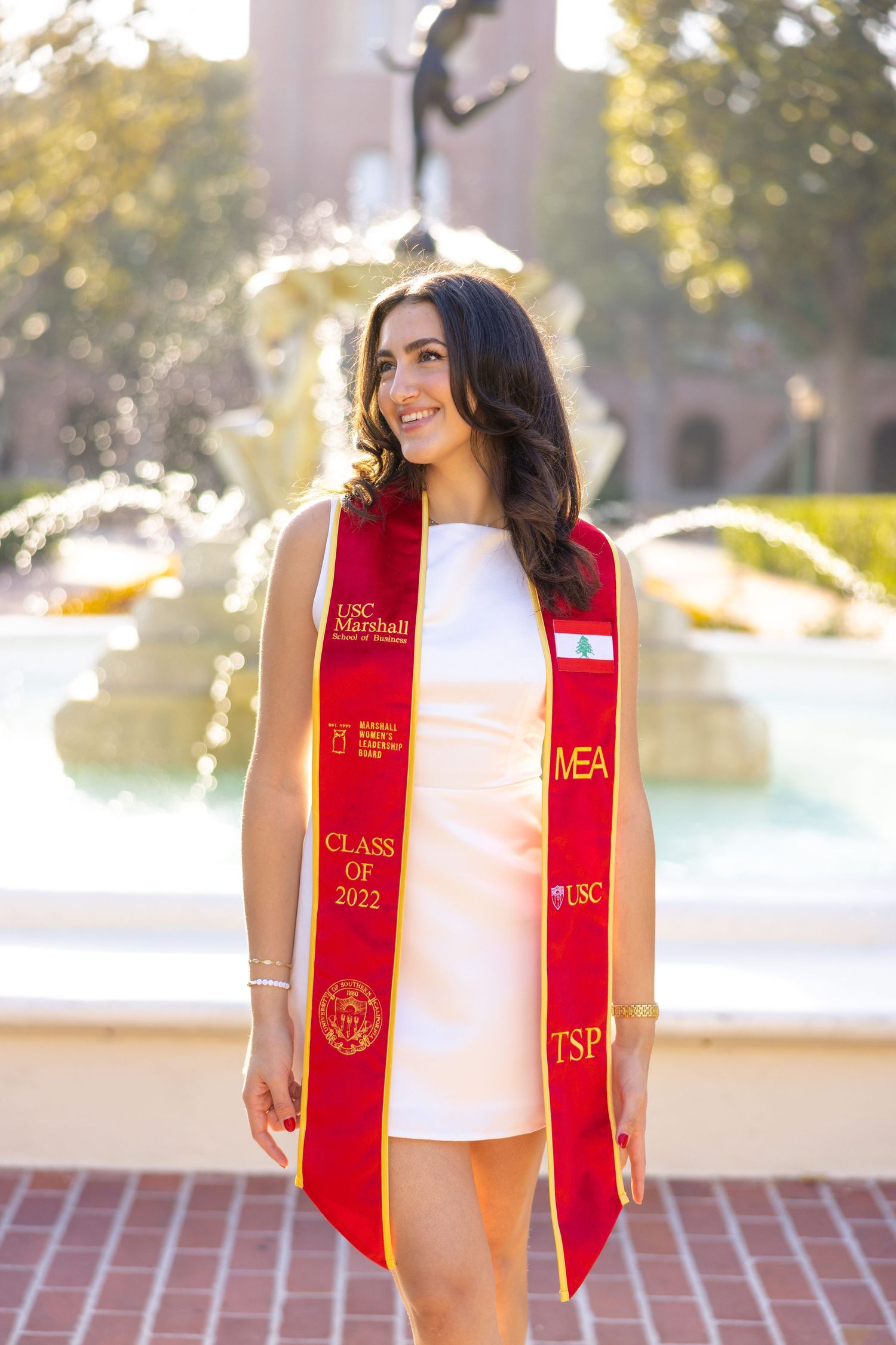 Graduate in white dress and red USC stole smiles in front of campus fountain.