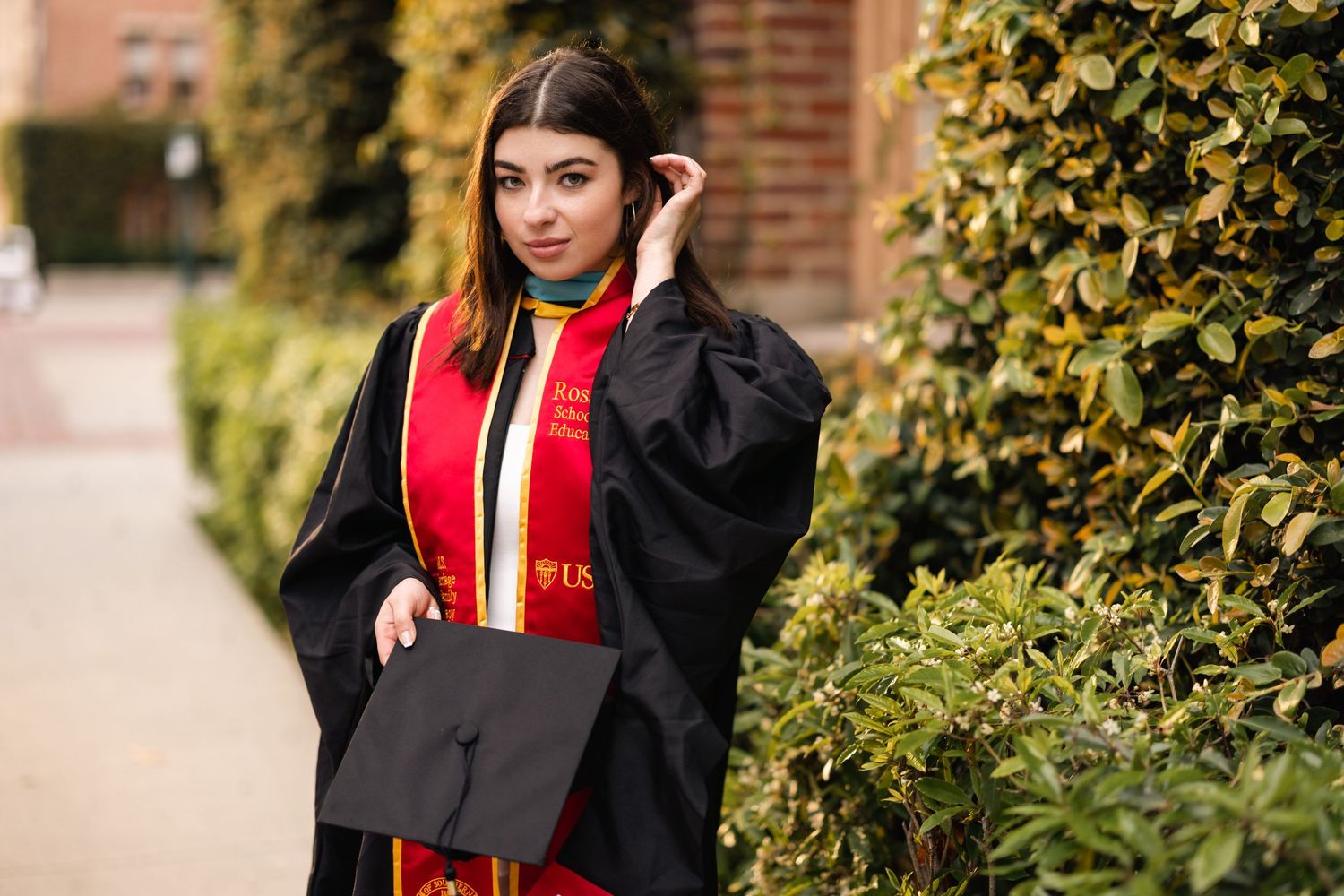 Graduate in black graduation gown and red sash posing by campus greenery.