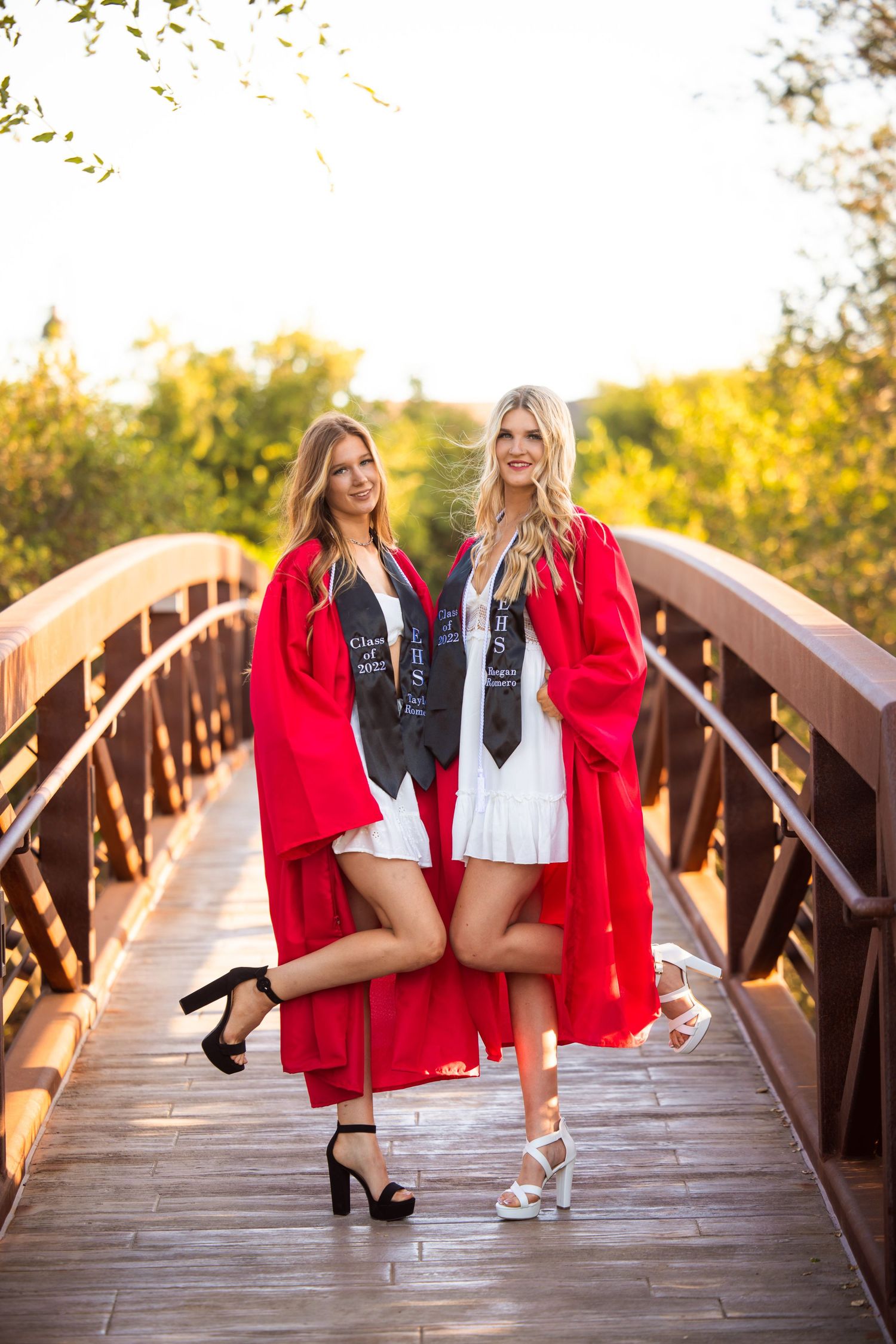 Two graduates in bright red robes pose playfully on a bridge during sunset.
