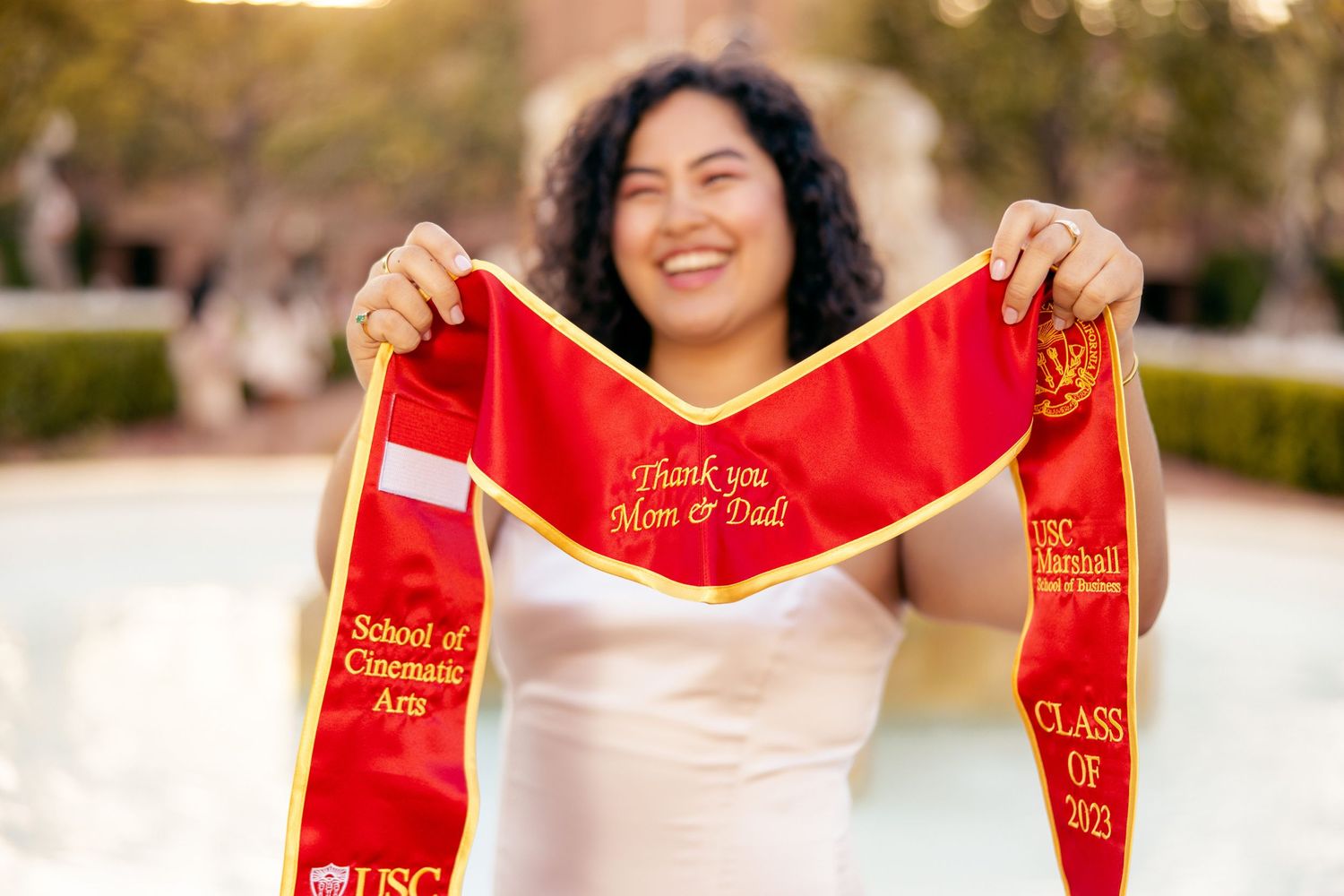 Graduate holding up red graduation sash with Class of 2023 text while smiling outdoors.