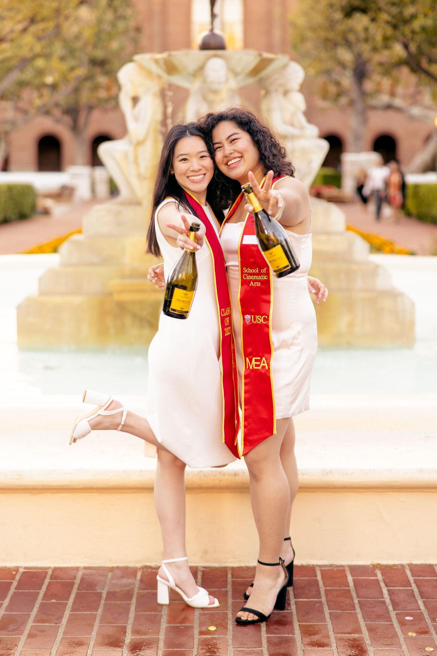 Two graduates in white dresses celebrating with champagne bottles by a fountain on campus.