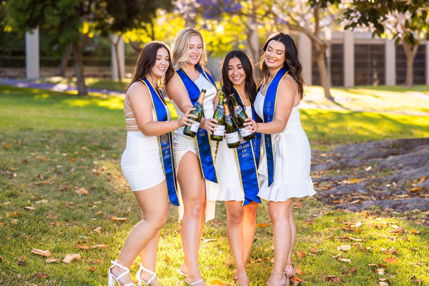 Group of graduates in white dresses and blue stoles celebrate with champagne bottles outdoors.