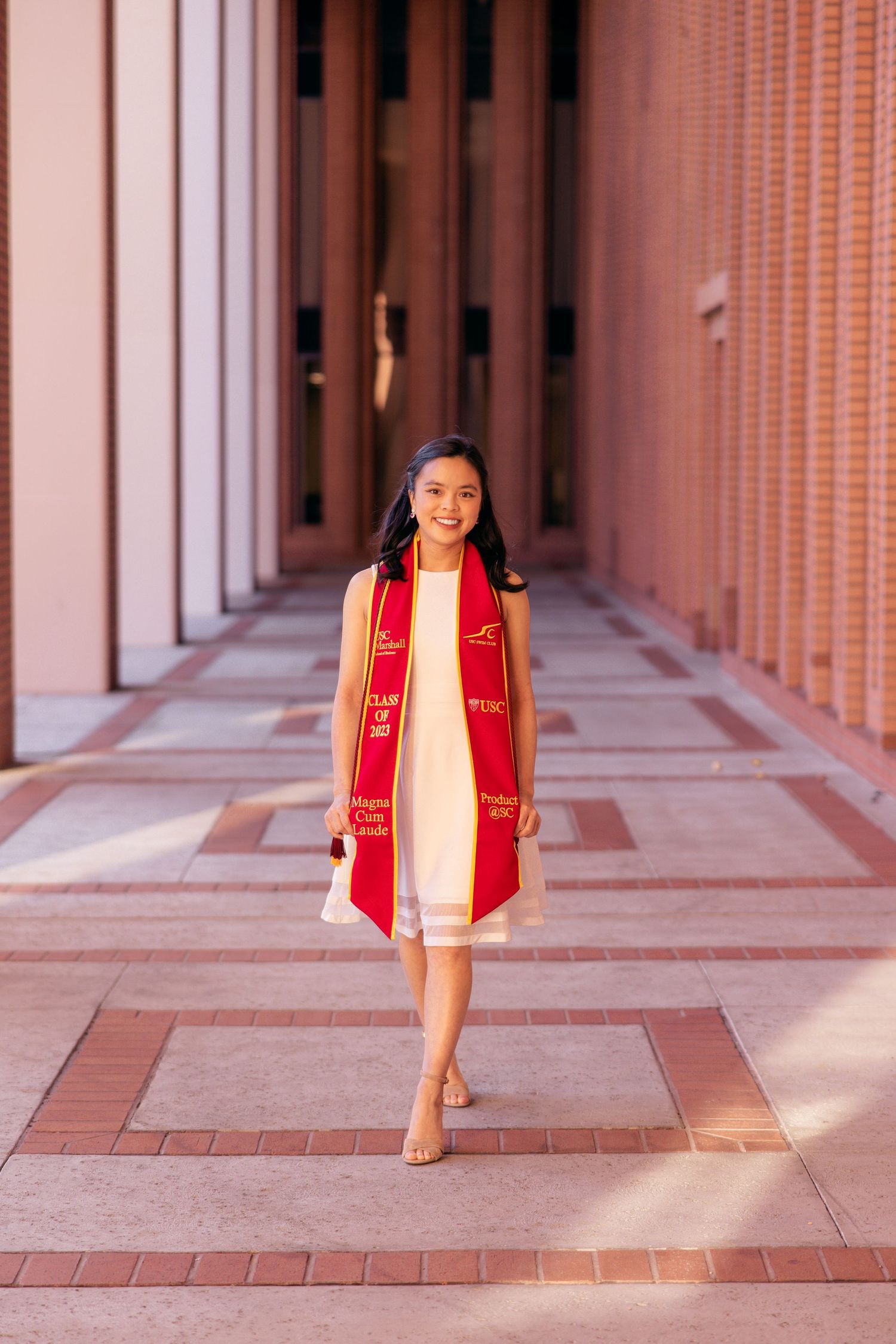 Graduate wearing white dress and red sash walking through arched campus hallway.