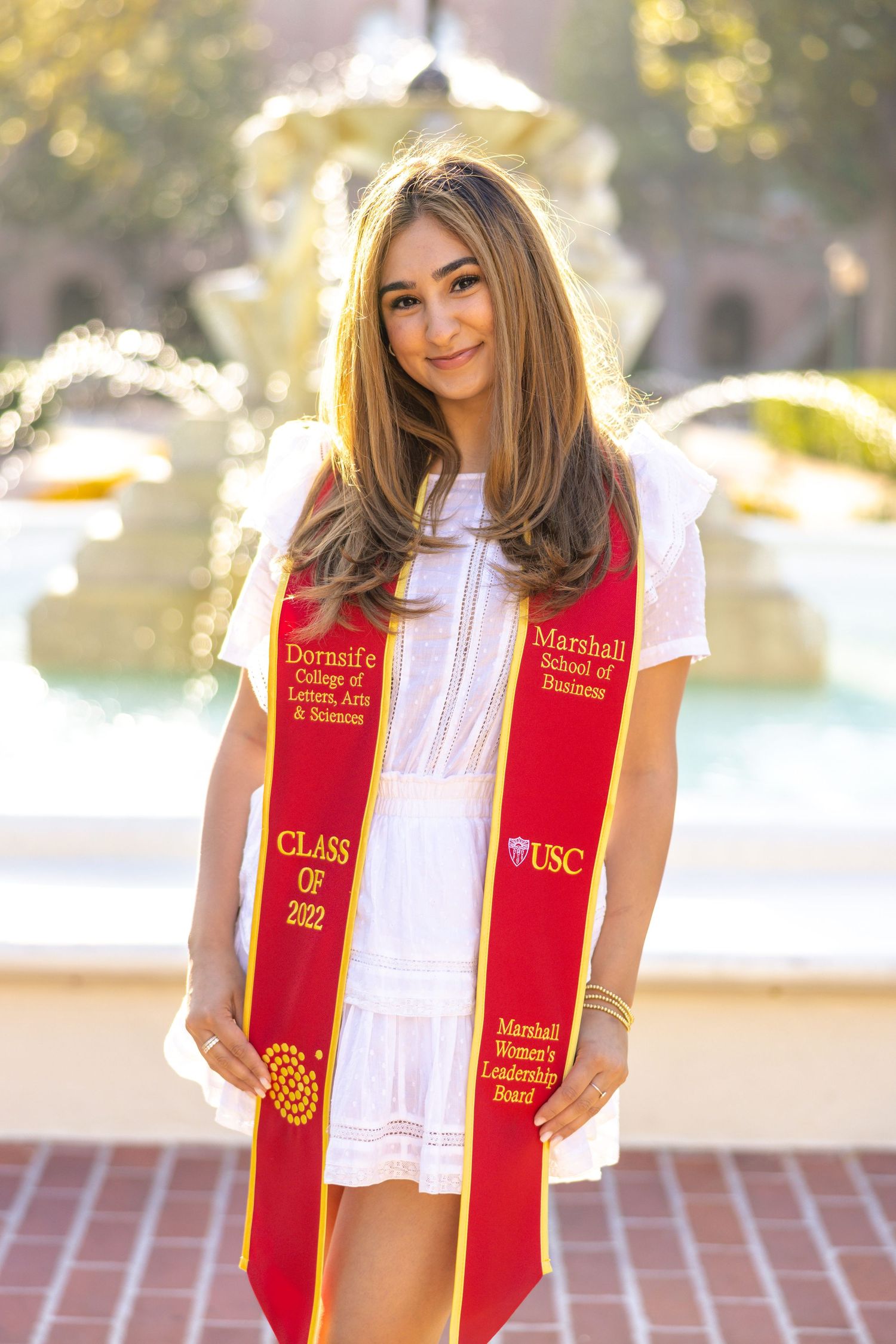 USC graduate wearing red and gold stole poses in front of campus fountain.