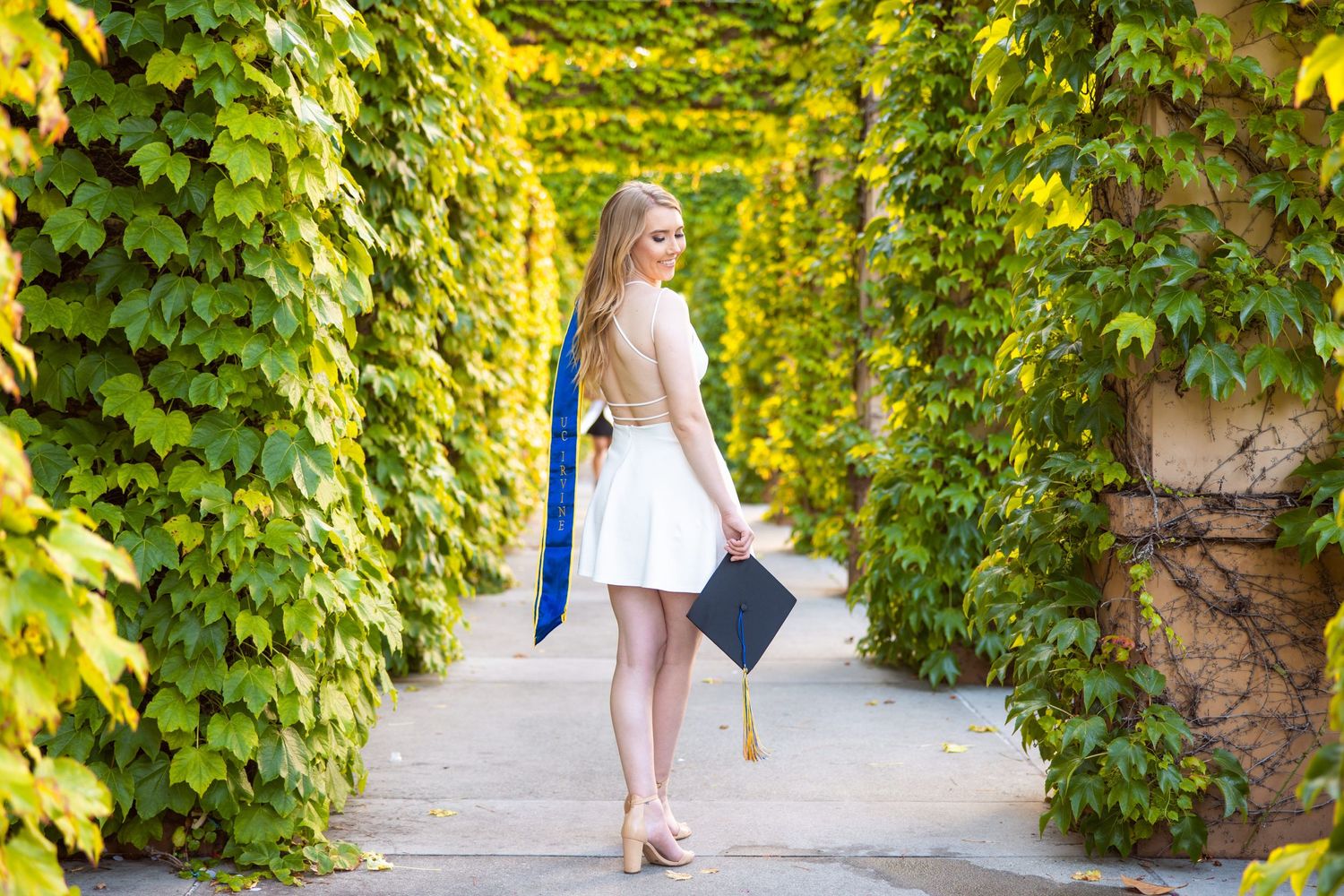 A graduate in a white dress poses in a garden pathway with ivy-covered walls.