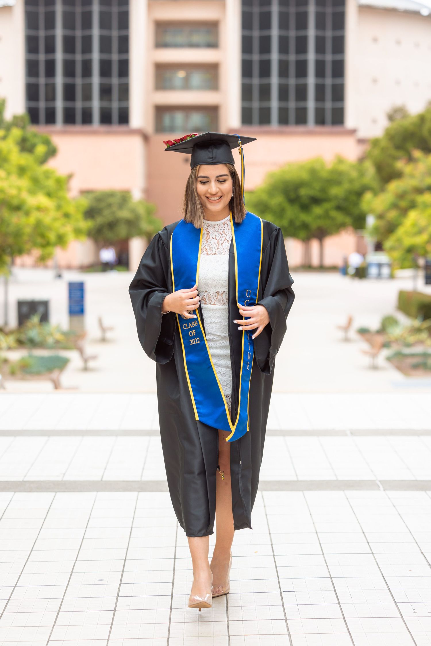 A graduate in cap and gown walks confidently across a university campus plaza.