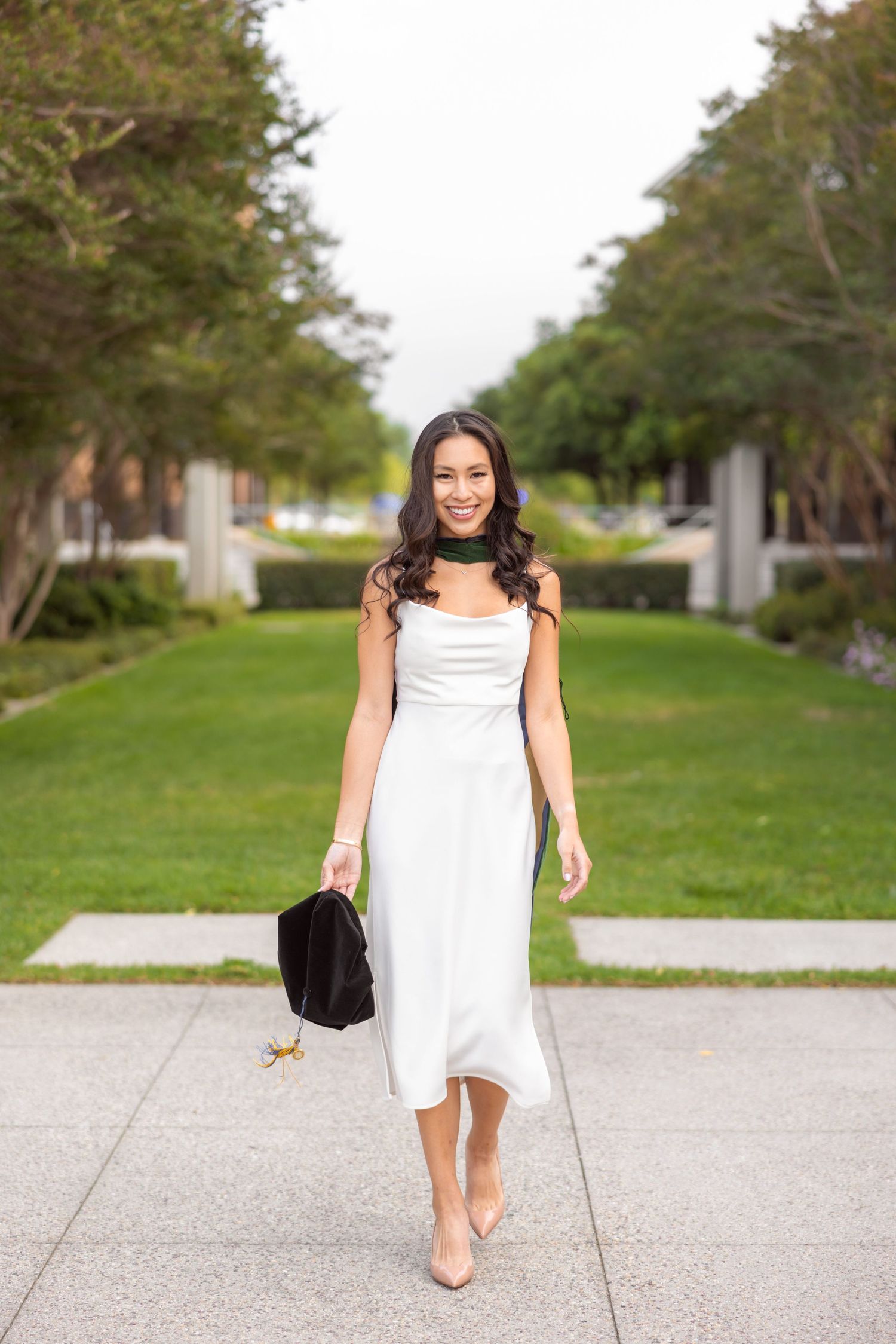 Elegant white midi dress styled with nude heels walking along a tree-lined path.