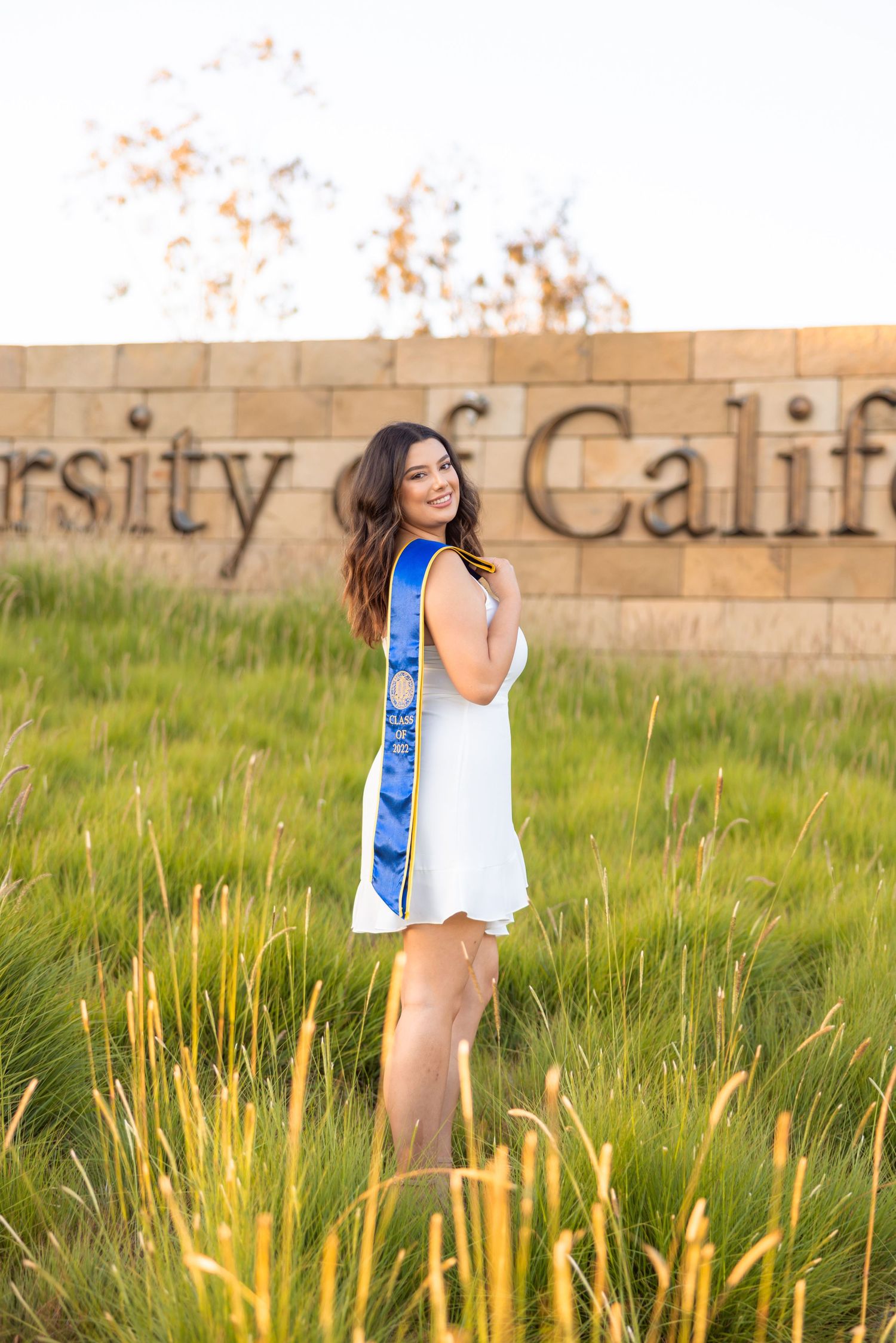 Graduate in white dress and blue stole poses in front of UC sign during golden hour.