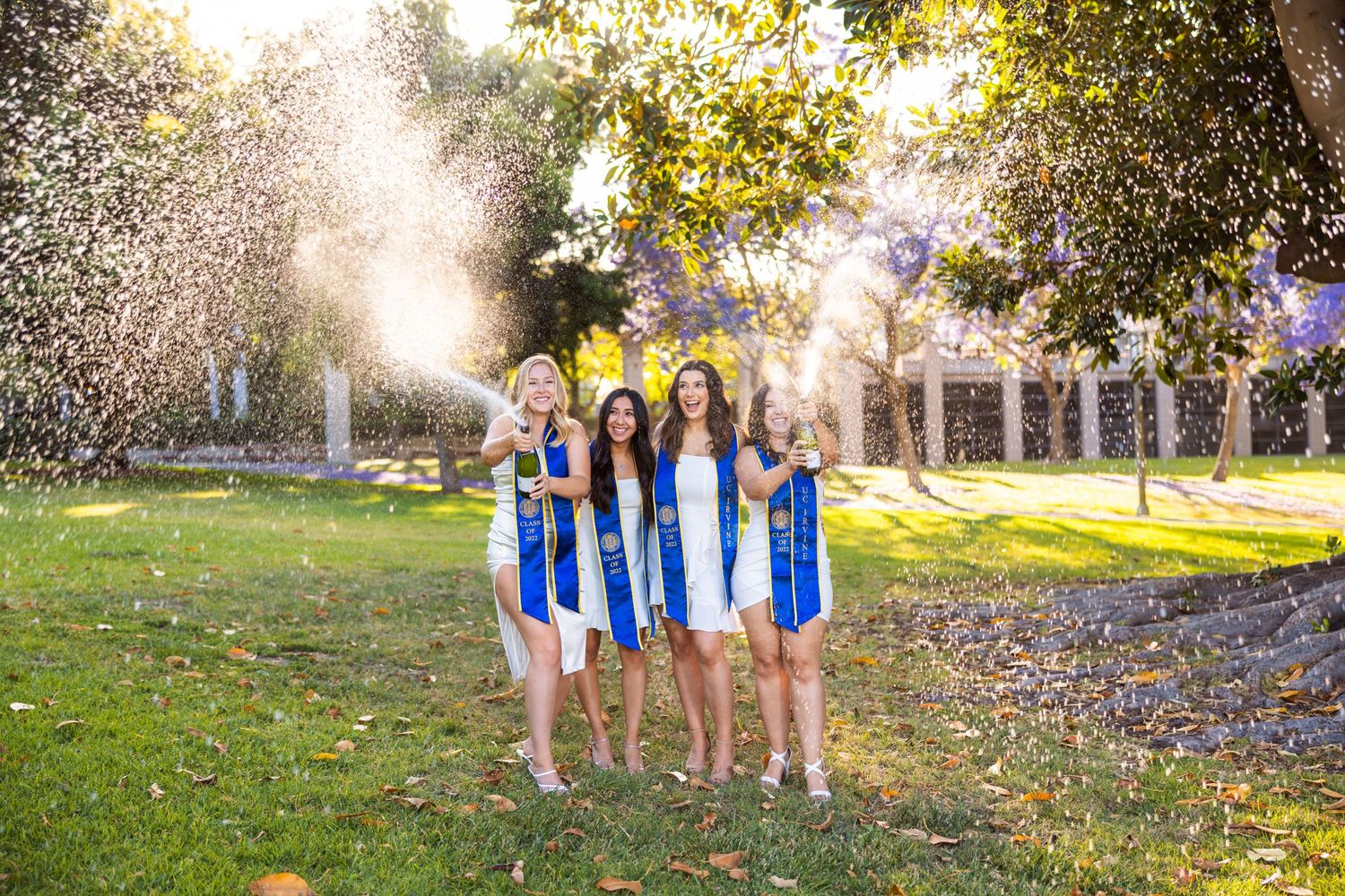 Graduates in white dresses celebrating with confetti toss under sunlit trees.