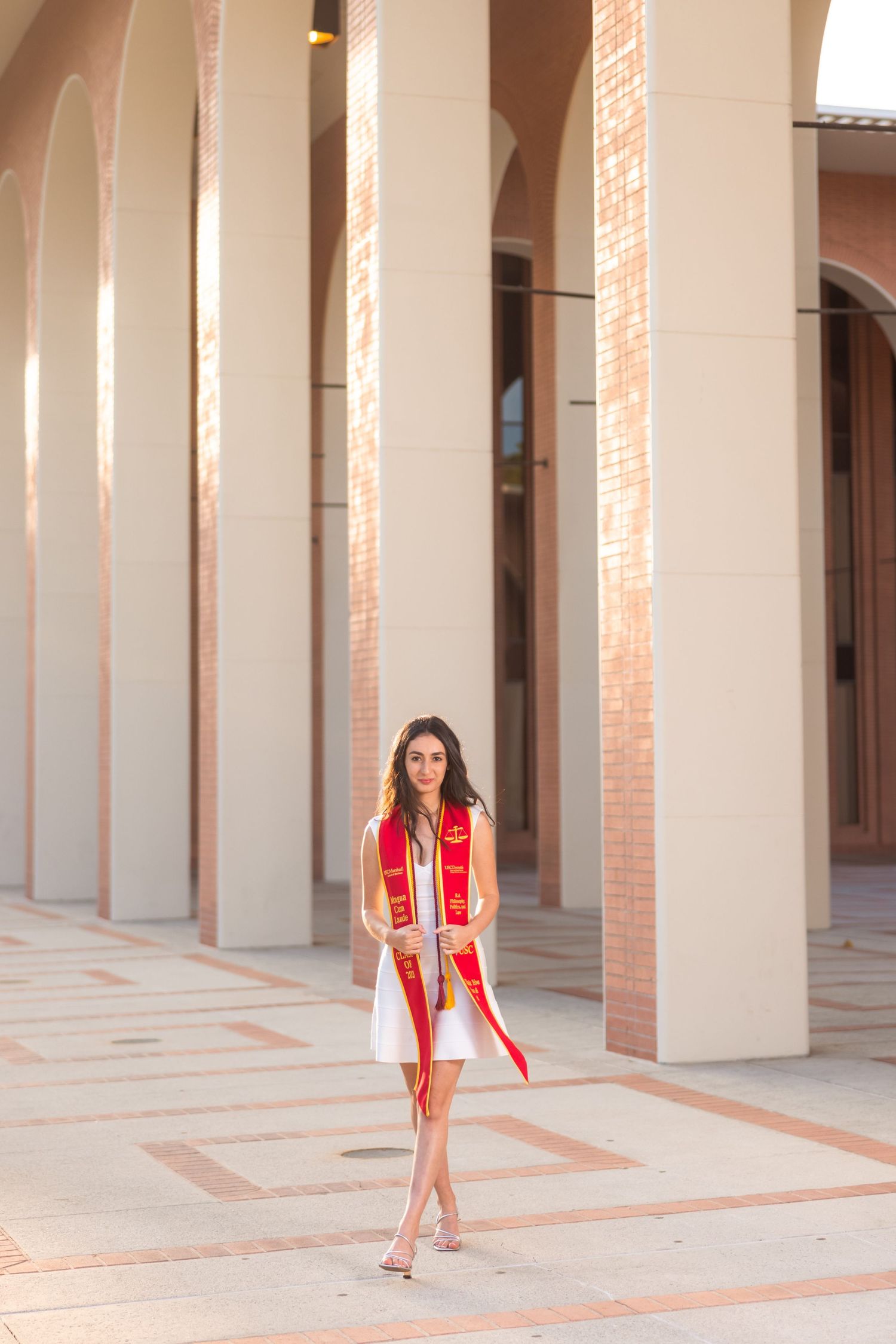 Stylish graduate in white dress and red stole poses between architectural columns.