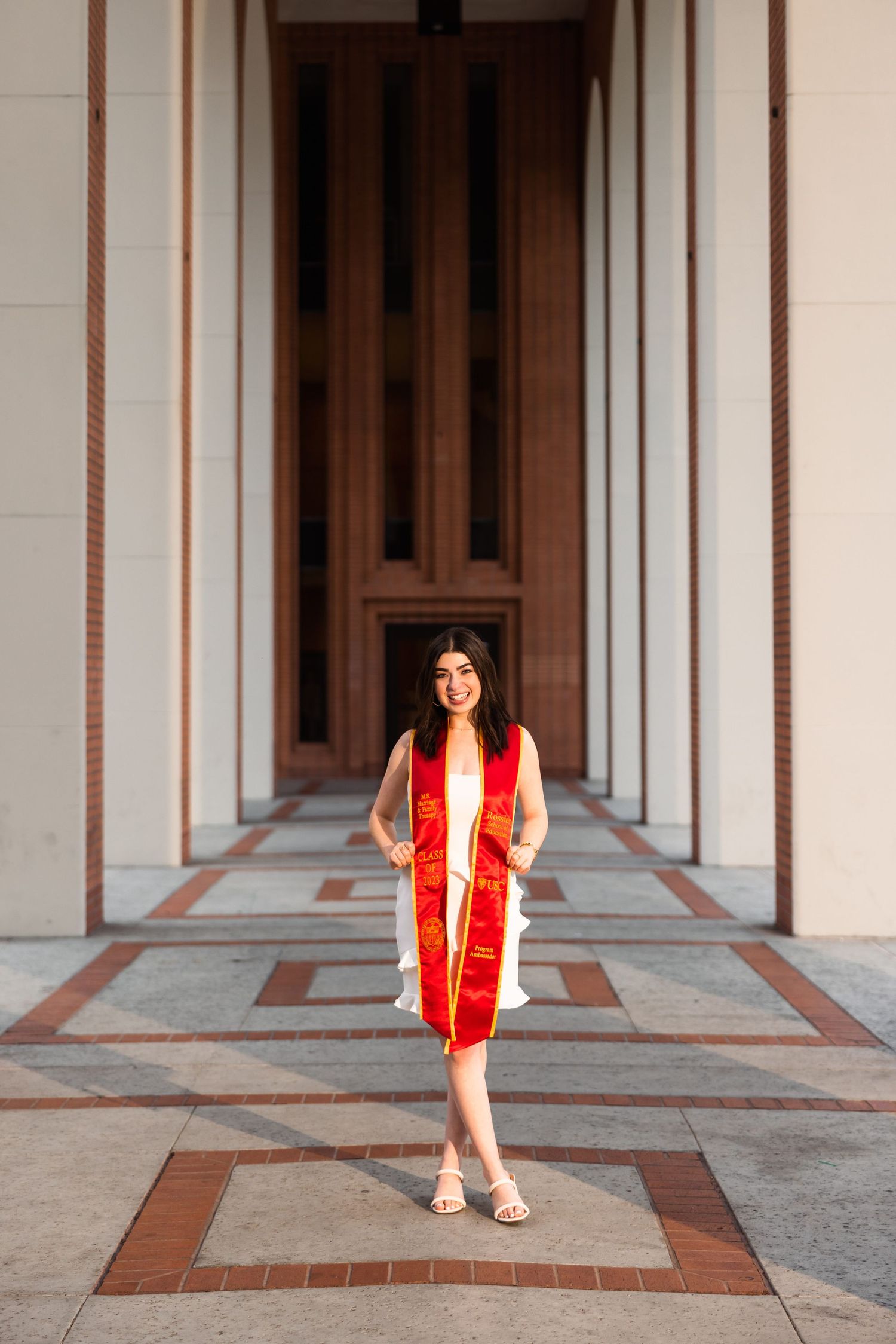 Graduate in white dress and red sash posing in architectural corridor.