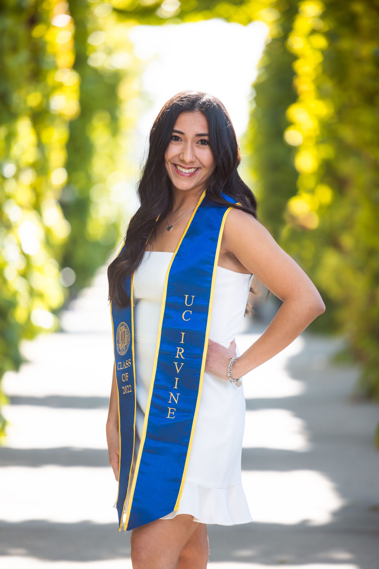 Graduate wearing UC Irvine blue sash poses in sunlit garden pathway.