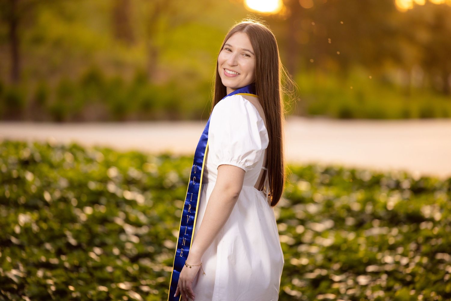 Graduate in white dress and blue stole poses in outdoor setting during golden hour.