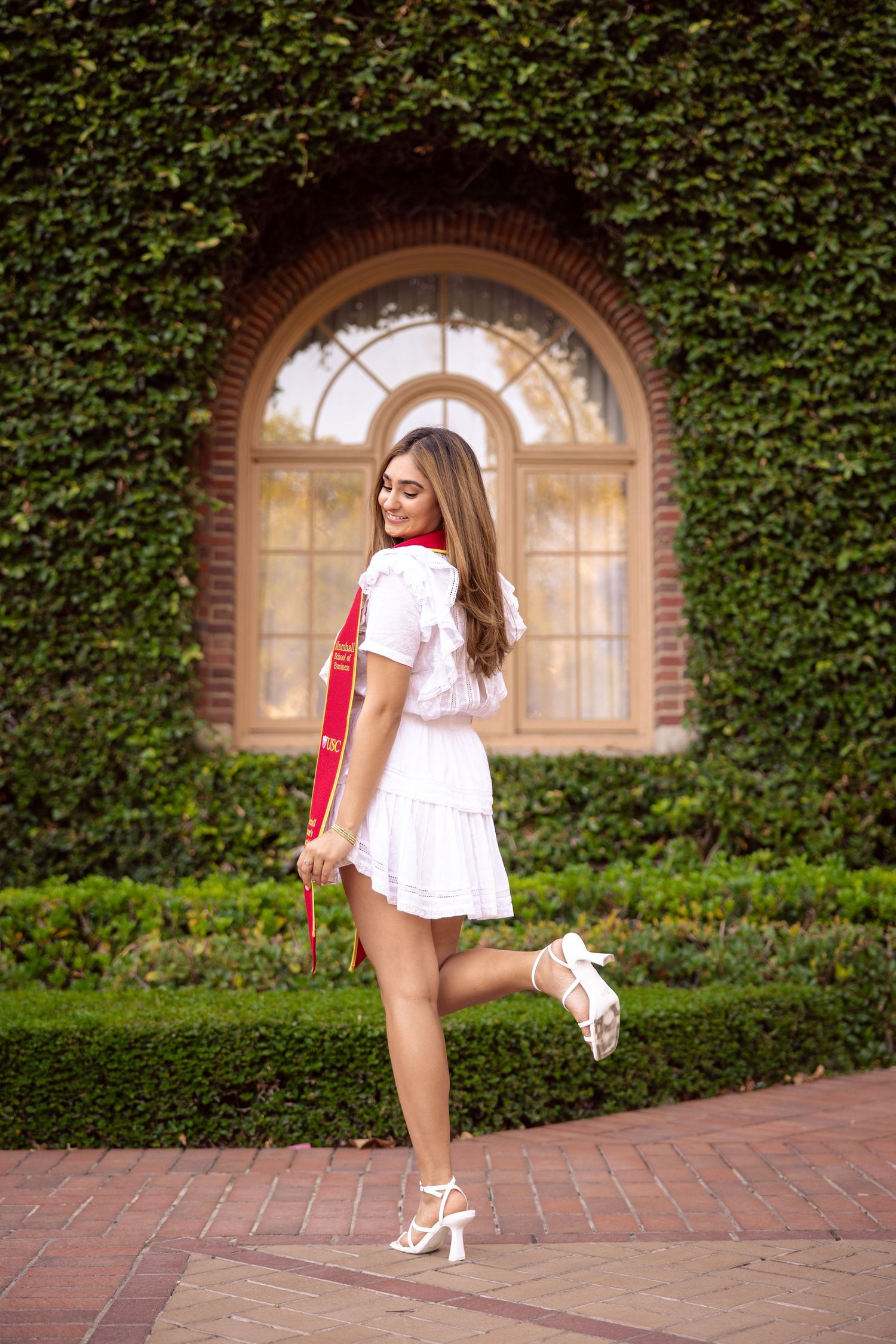 Graduate in white dress and red stole poses playfully by ivy-covered archway.