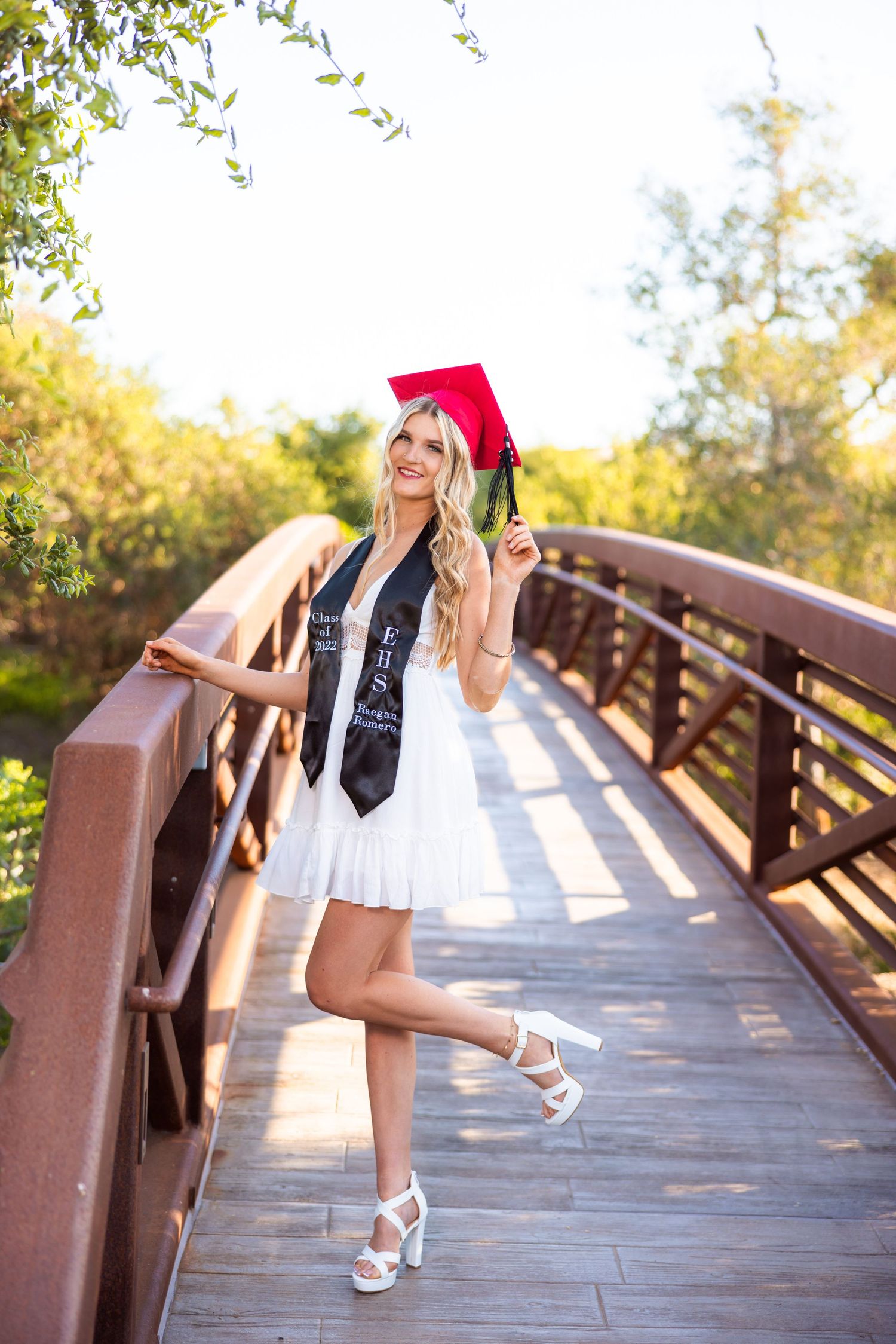Graduate in white dress and red cap poses on wooden bridge during golden hour.