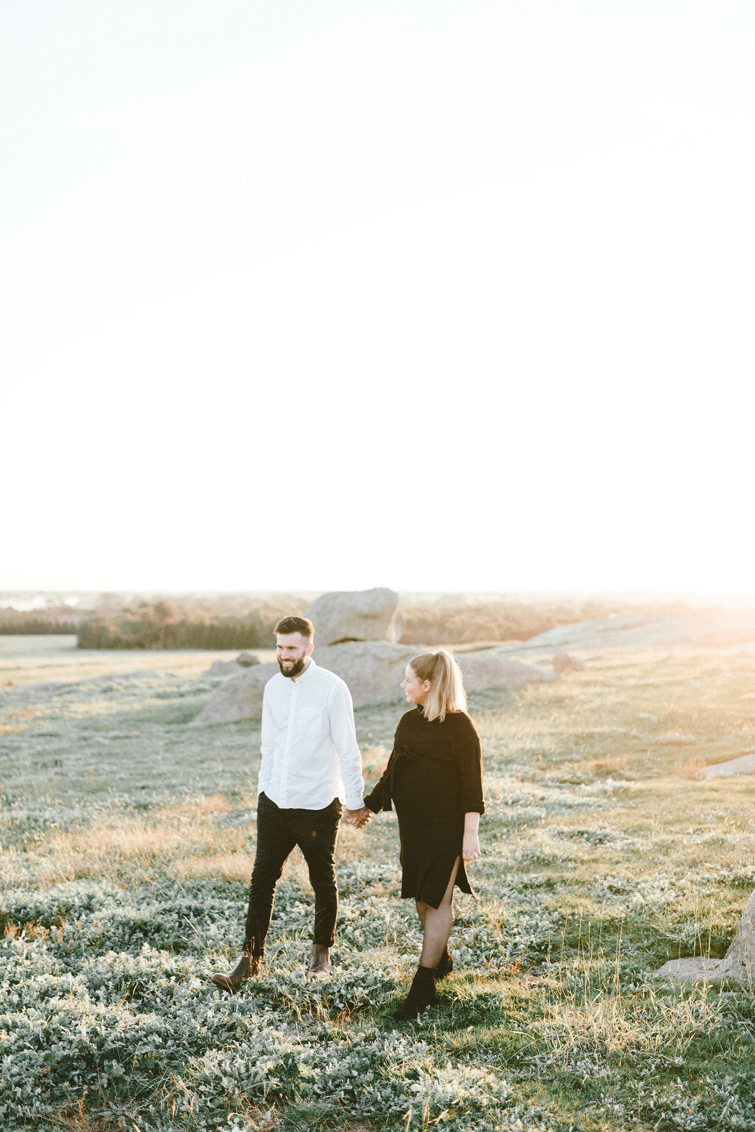 A couple walks through a grassy meadow at sunset with rolling hills in the background.
