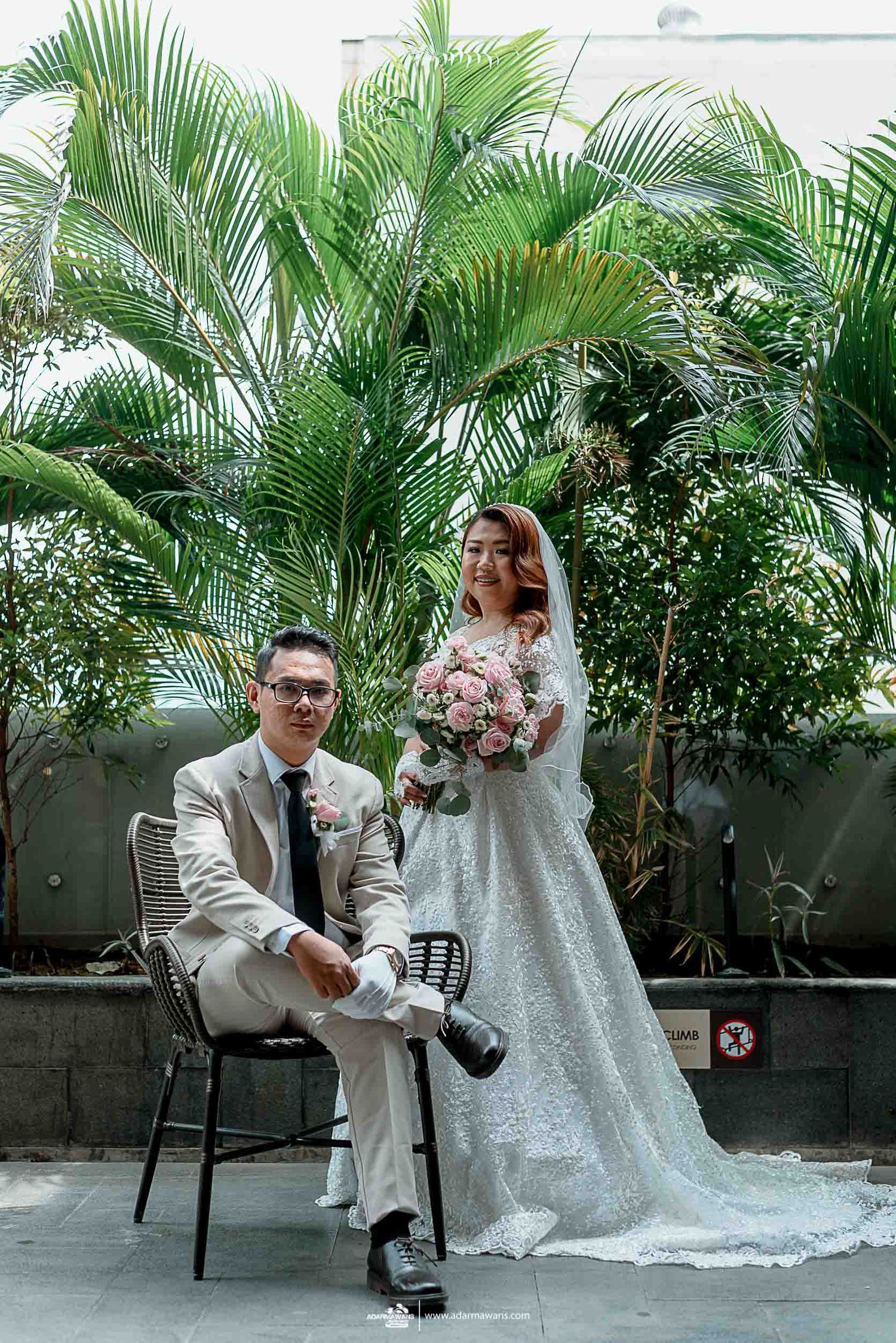 Wedding couple poses among tropical palm plants, with bride holding pink bouquet.