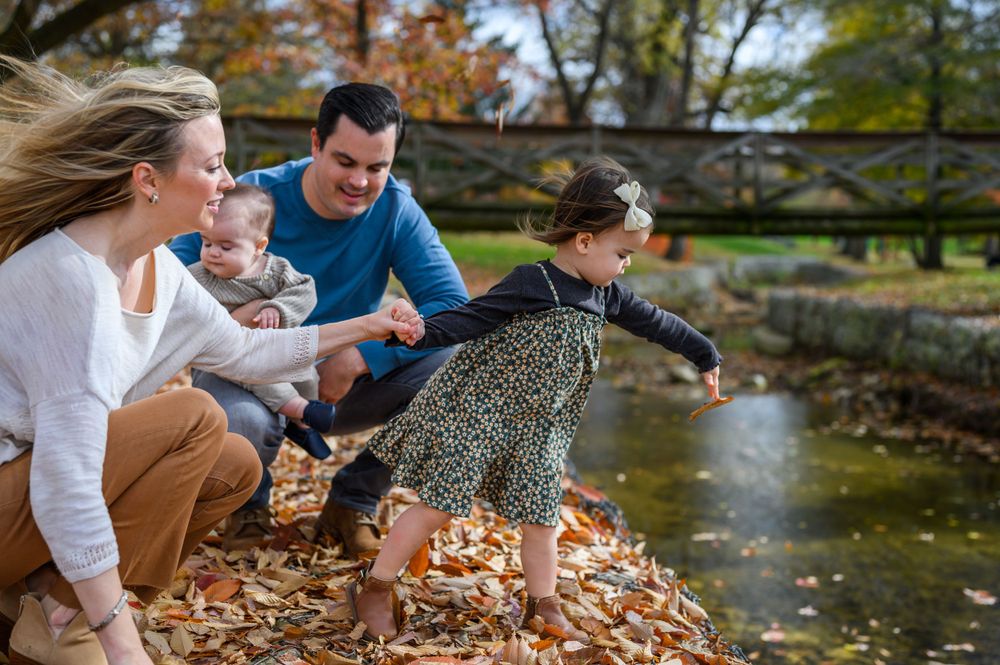 Family playing in fallen autumn leaves by a pond on a sunny day.