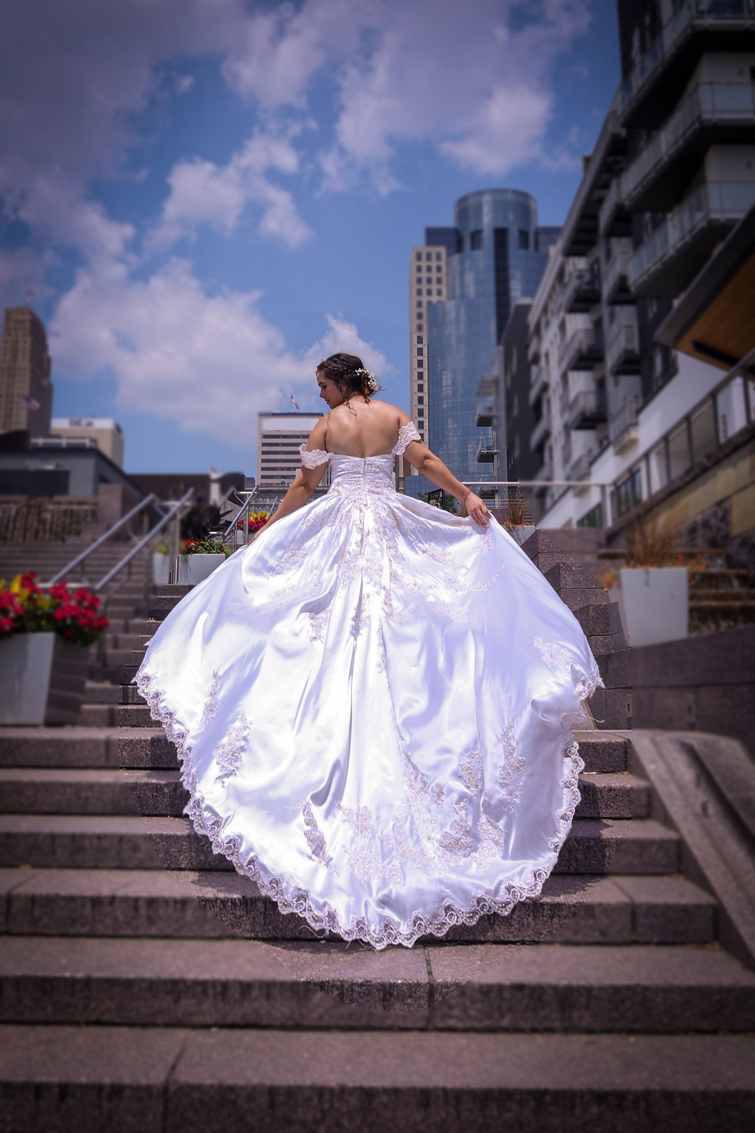 A bride in a lace wedding gown with flowing train ascends concrete steps in an urban setting.