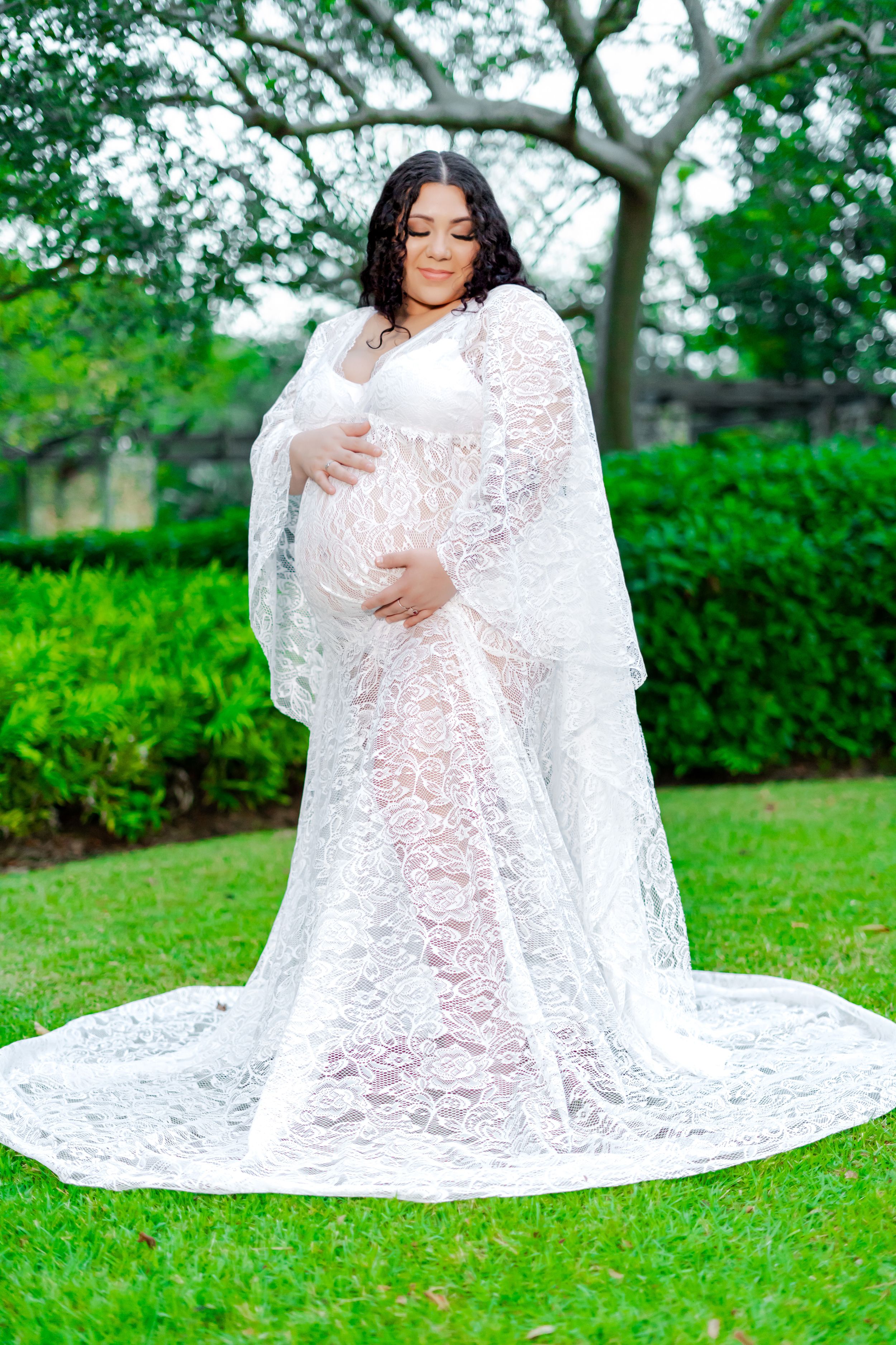 An expectant mother poses in a flowing white lace maternity gown with a long train in a lush garden setting.