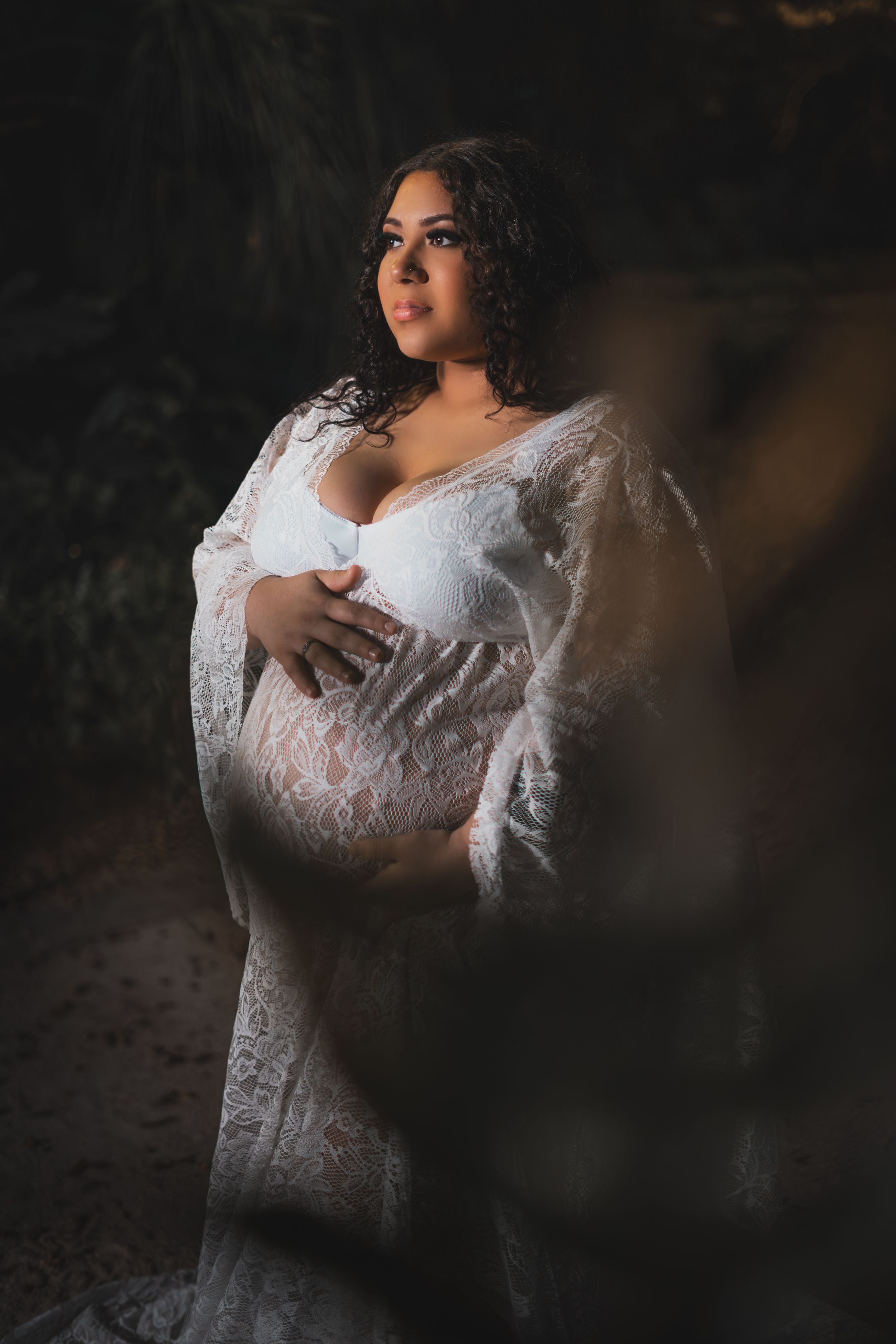 An expectant mother in a white lace gown poses for a dramatic maternity portrait.