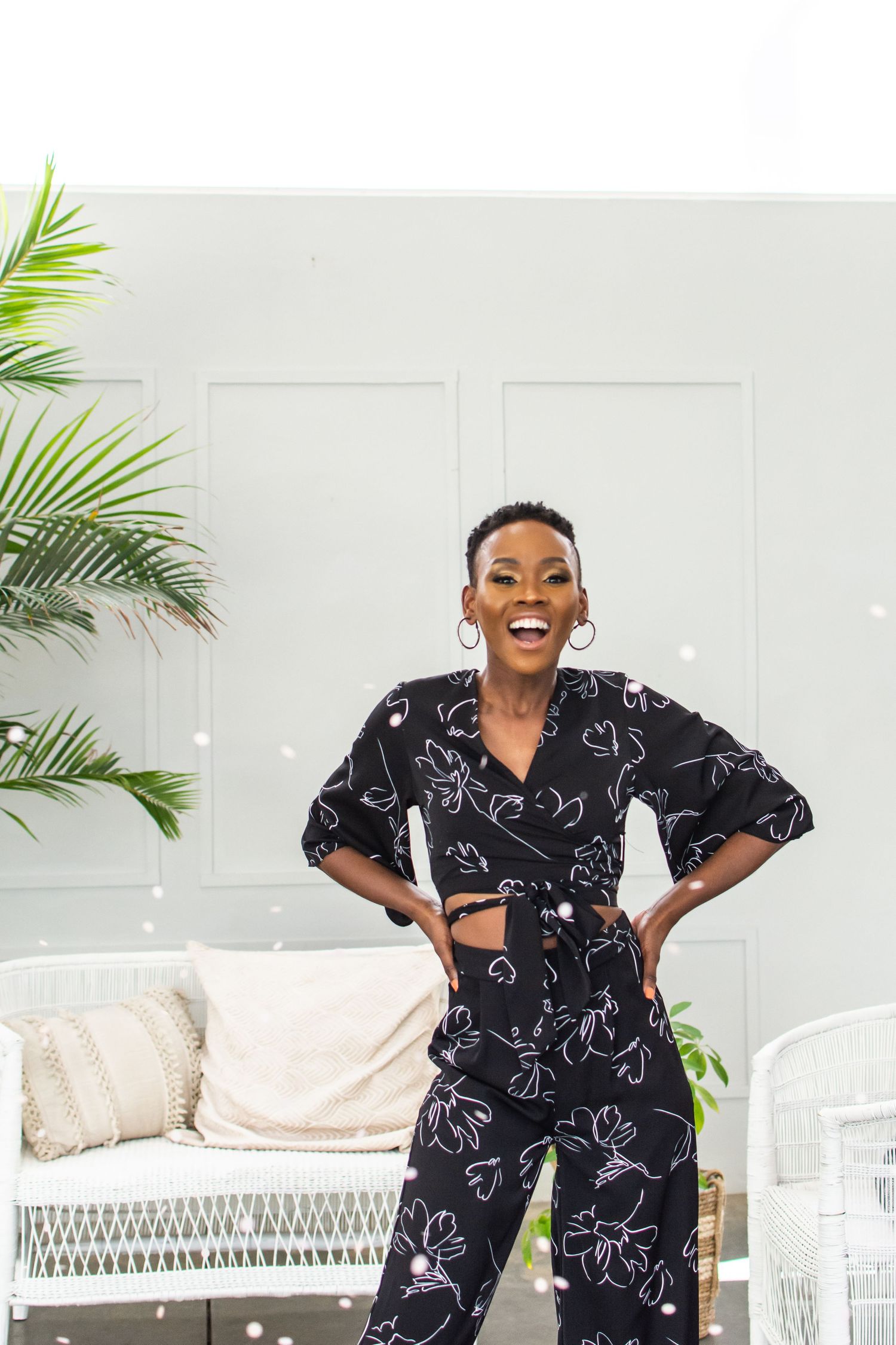 Dynamic pose in black floral two-piece outfit against white wicker furniture and tropical backdrop.