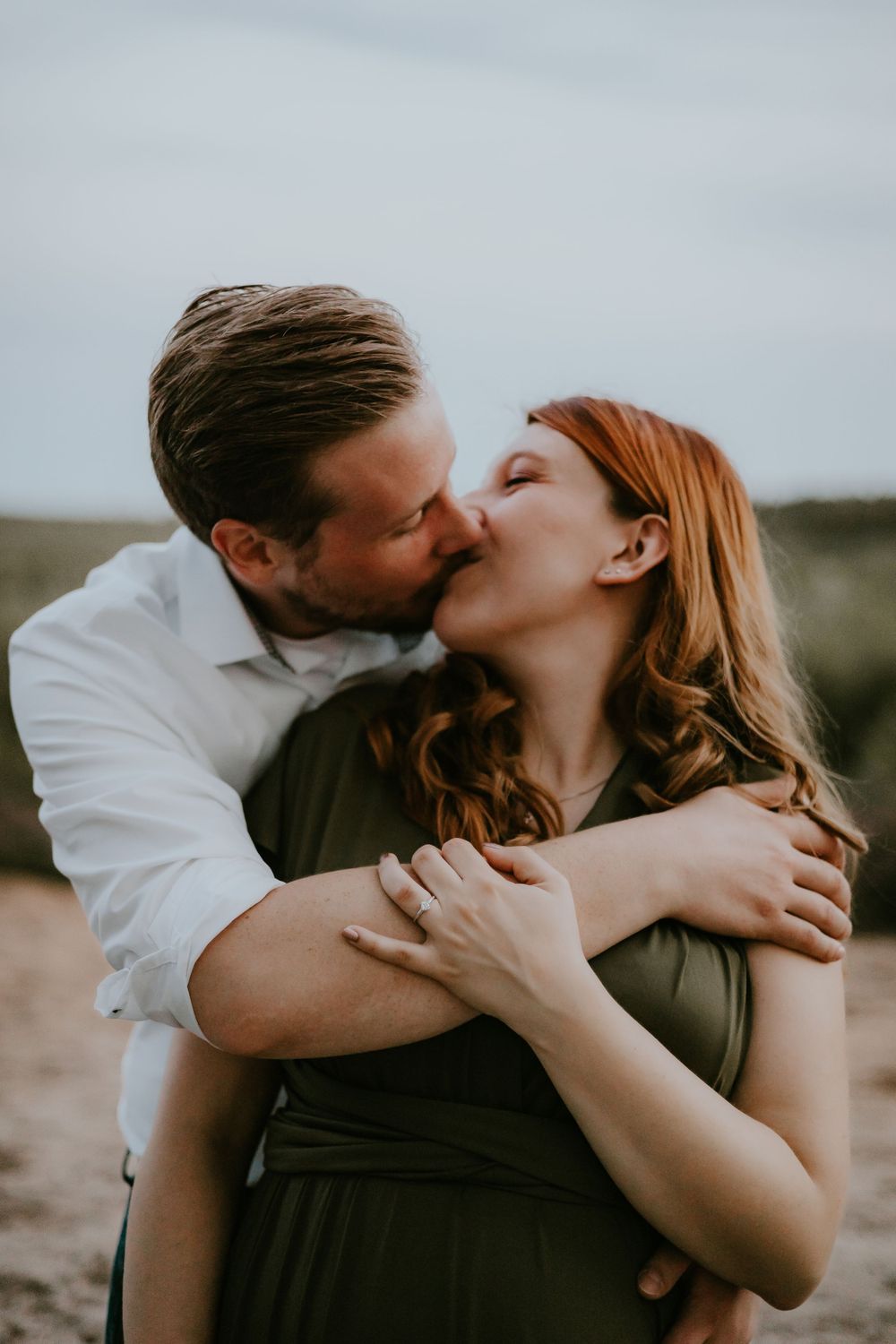 A romantic couple shares a tender kiss outdoors, with the woman wearing an olive green dress and the man in a white shirt.