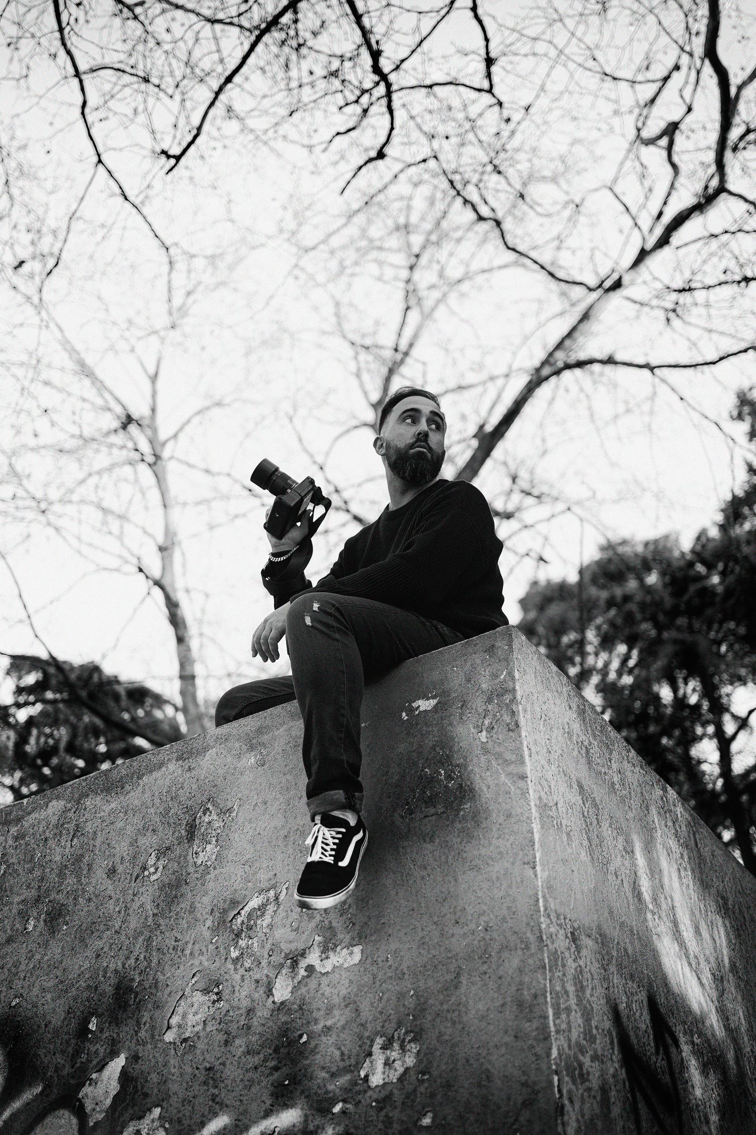Black and white photo of photographer sitting on large rock under bare tree branches.