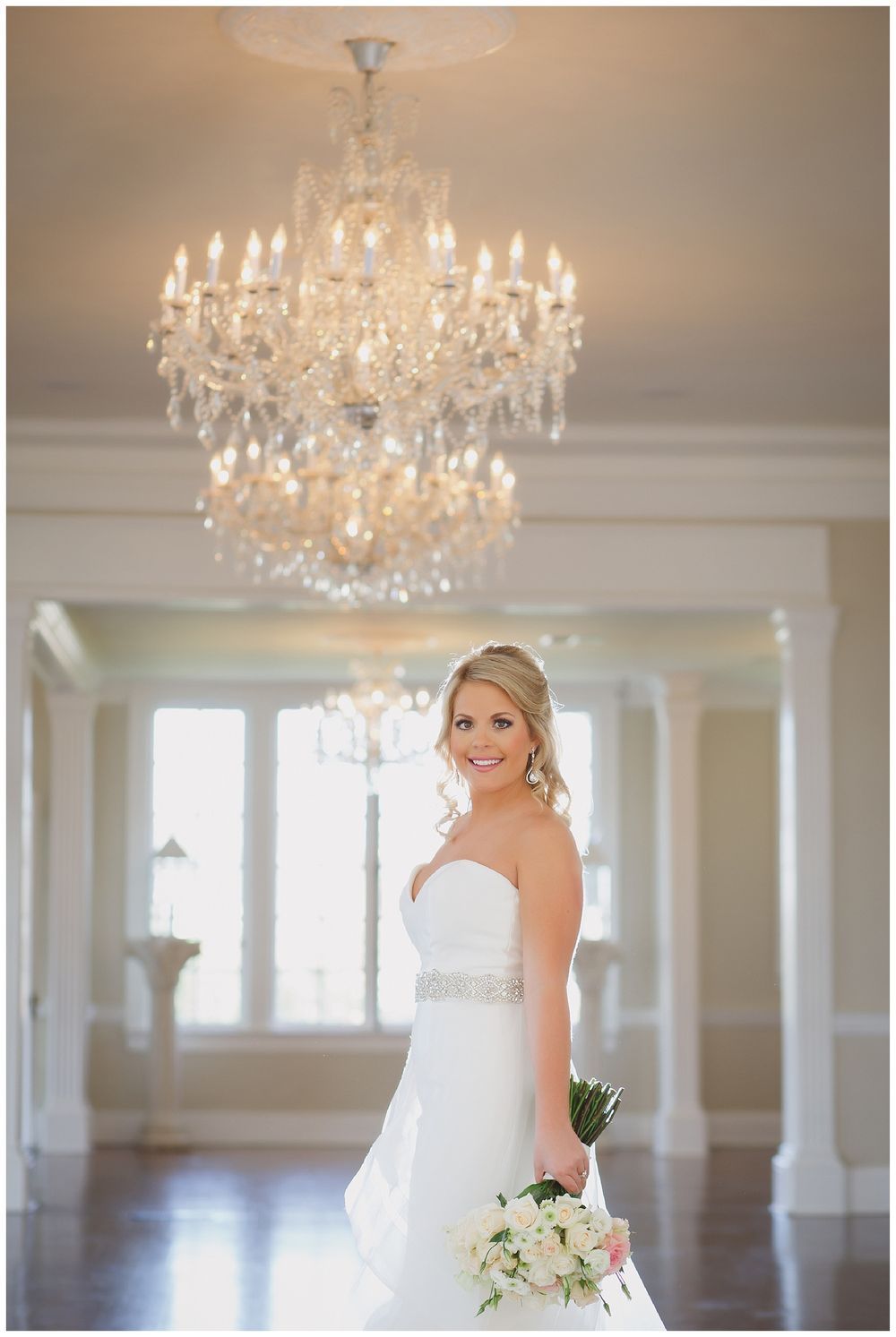 Elegant bridal portrait under crystal chandelier in luxurious white room.