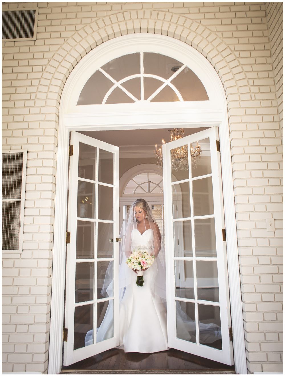 Bride stands in elegant arched doorway of white brick building holding bouquet.