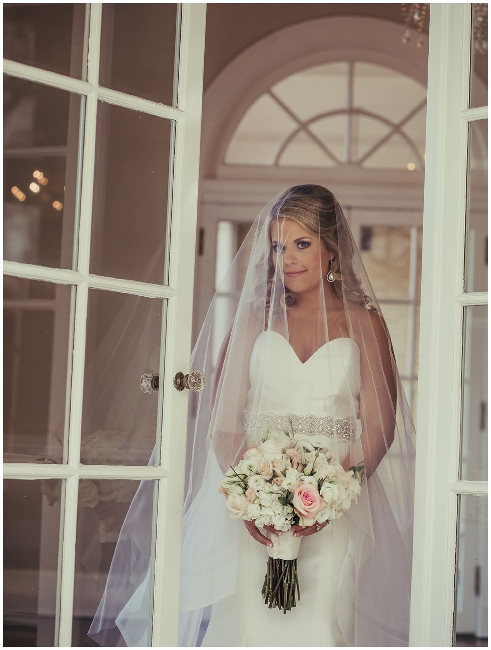 Elegant bride in flowing white dress and veil holding a blush and white floral bouquet near French doors.