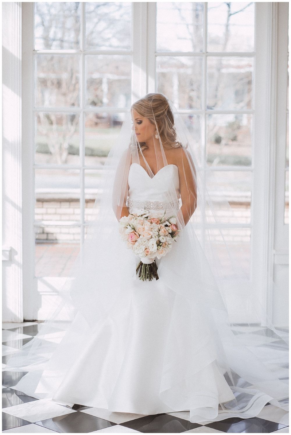 Bride in classic ballgown poses in sunlit room with checkered floors and pristine white windows.