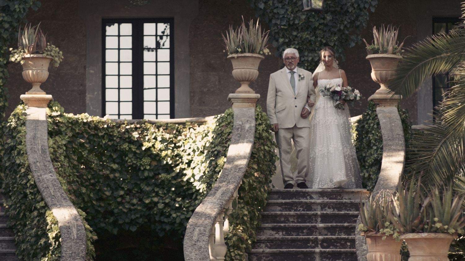 Elegant wedding couple descends ornate stone staircase decorated with ivy and potted plants.