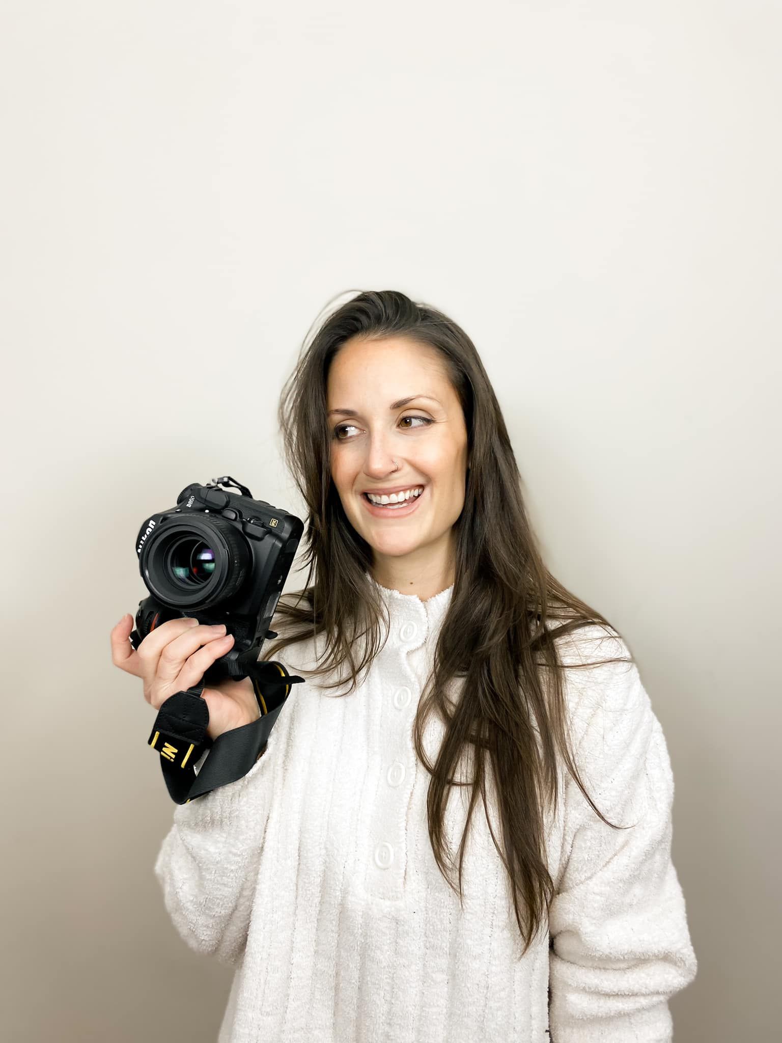 Professional photographer in white sweater holding DSLR camera against neutral background.