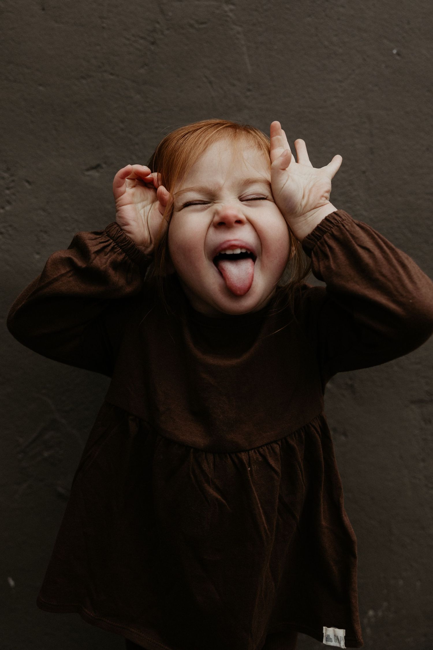 Playful redhead toddler girl in brown dress sticking out tongue and making funny face against dark wall. Miranda Photo & Co